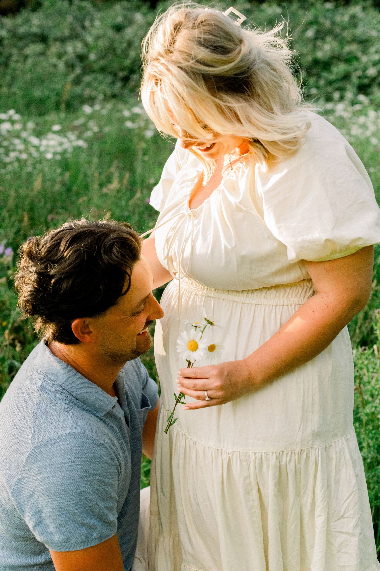 A man kneels before a pregnant woman in a white dress, holding daisies near her belly in a sunny green field.