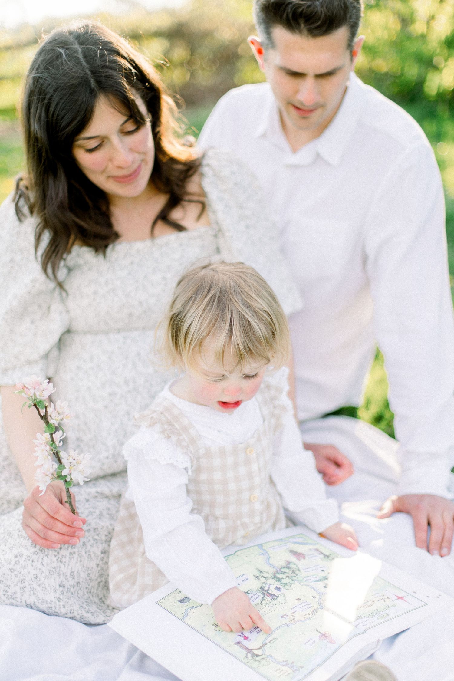 A young family enjoys a sunny outdoor picnic, with their toddler exploring a colorful picture book together.