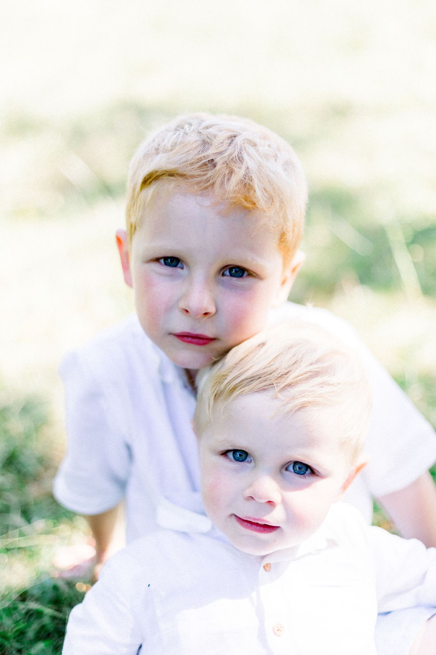 Two blonde blue-eyed brothers in white shirts pose together outdoors in soft natural light.