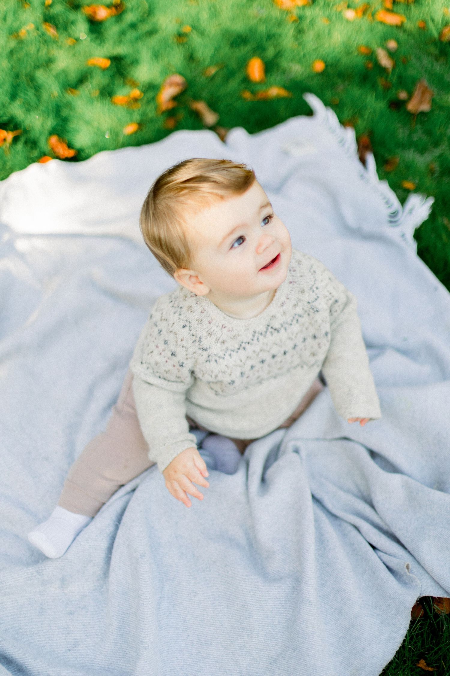 A toddler in grey sweater crawls on blue blanket with green grass and orange leaves visible.
