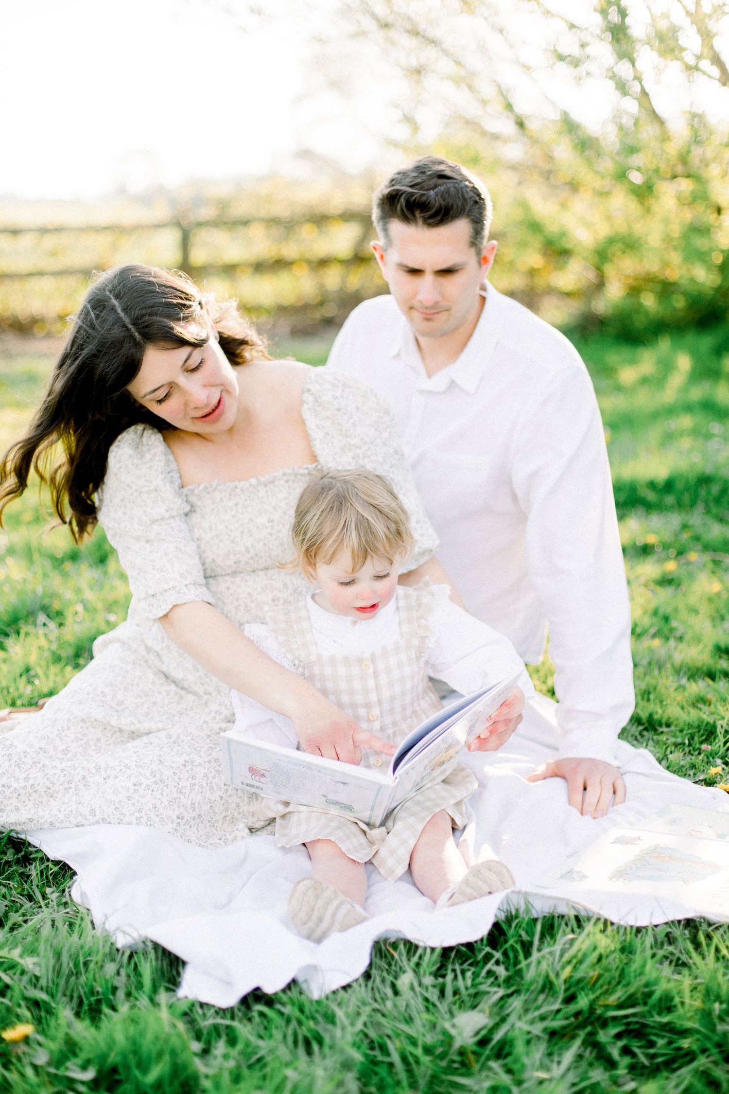 A family reading together outdoors on a white blanket during a sunny afternoon.