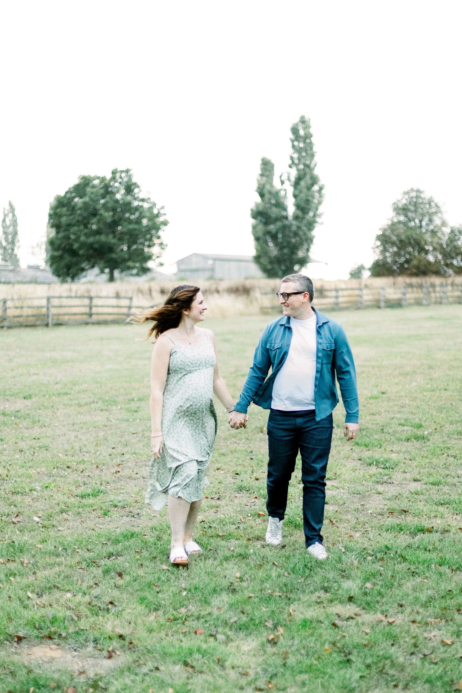 A couple holds hands walking through a green field, woman in floral dress, man in denim jacket, with trees and fence behind.