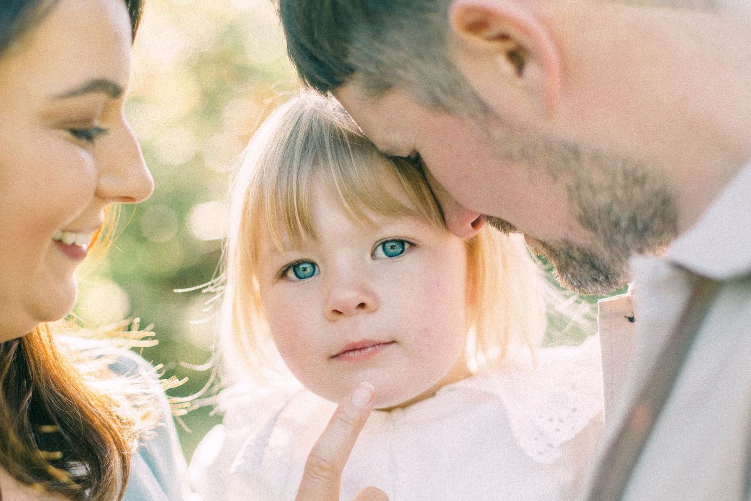 Blue-eyed toddler girl in a white lace dress gazes at the camera while her smiling mother and bearded father lean in close.