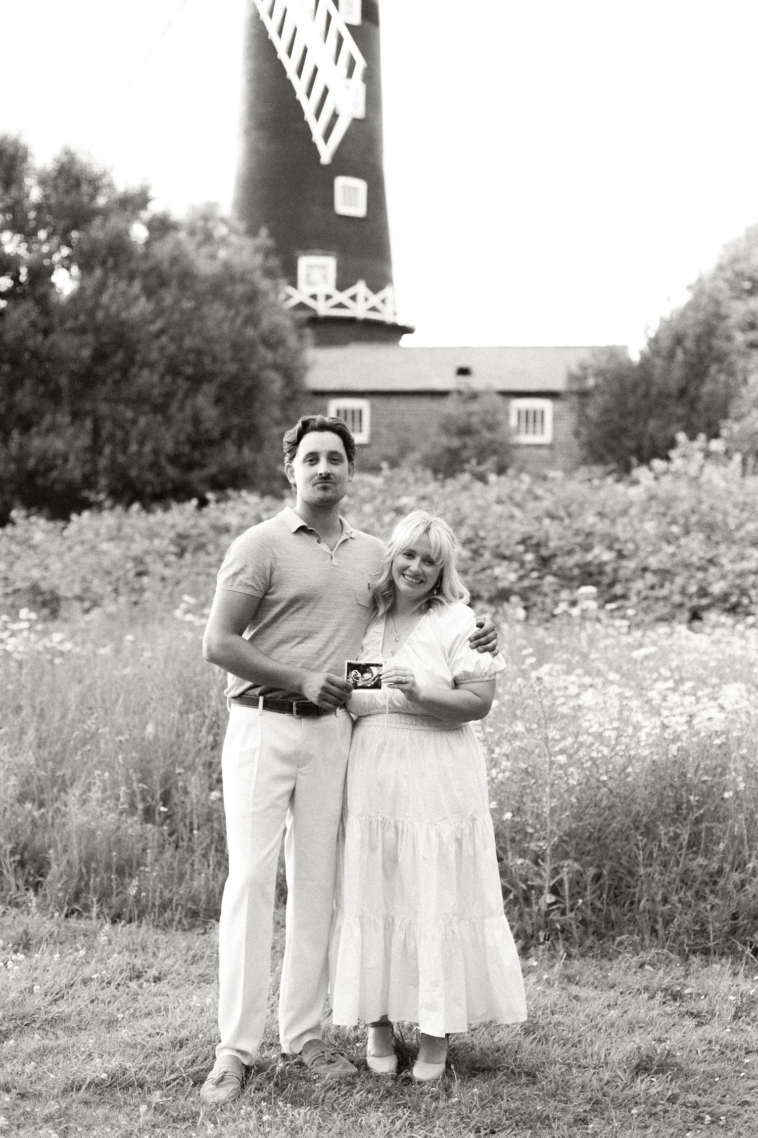 Black and white photo of a couple holding an ultrasound scan in front of a windmill.