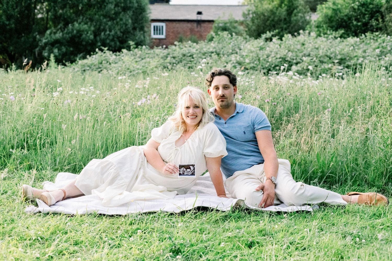 Smiling couple sitting on a white blanket in a grassy meadow, the woman holding an ultrasound photo in a sweet pregnancy announcement portra