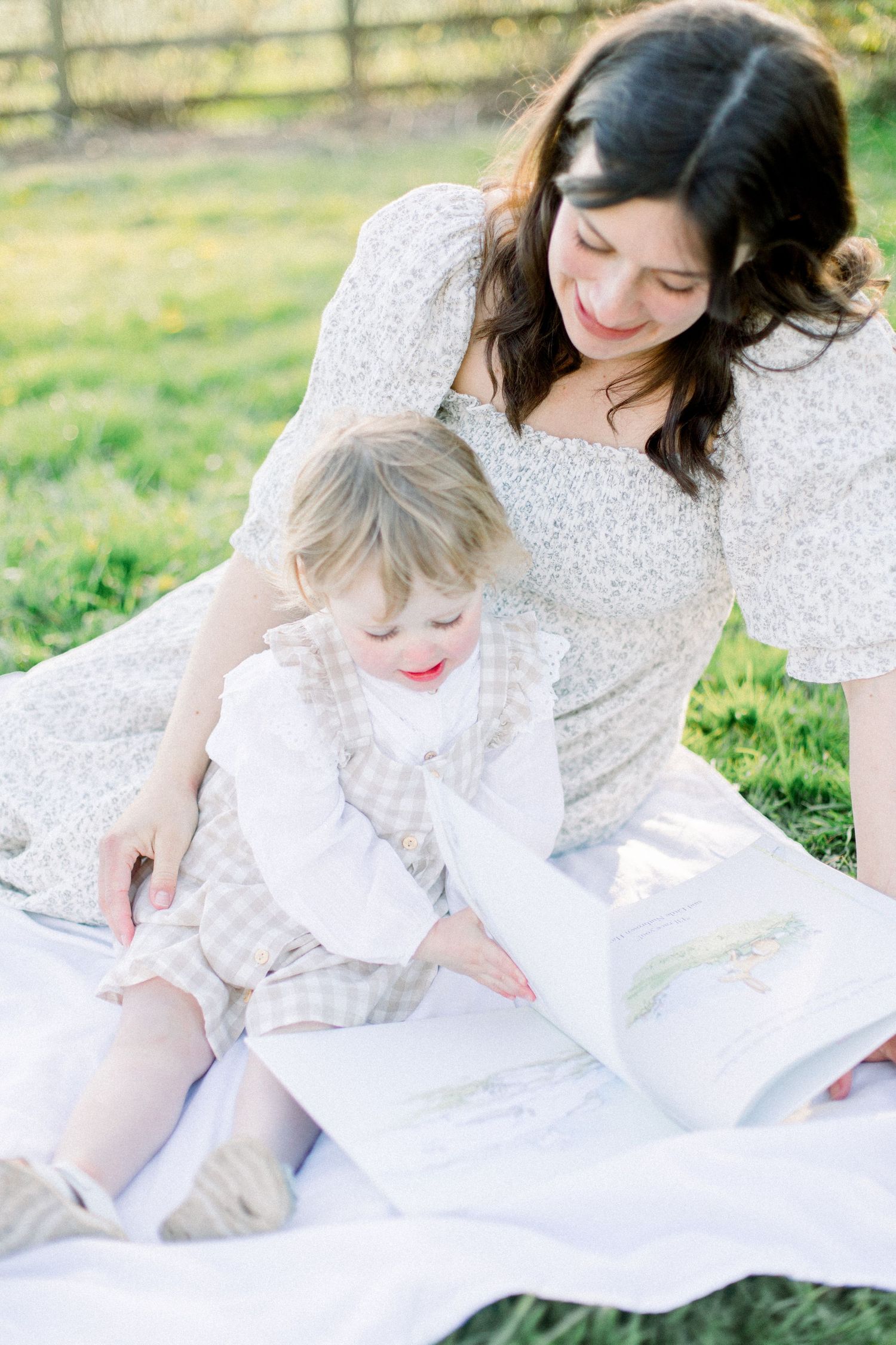 A mother and toddler sit on a white blanket outdoors, reading a picture book together in soft natural light.