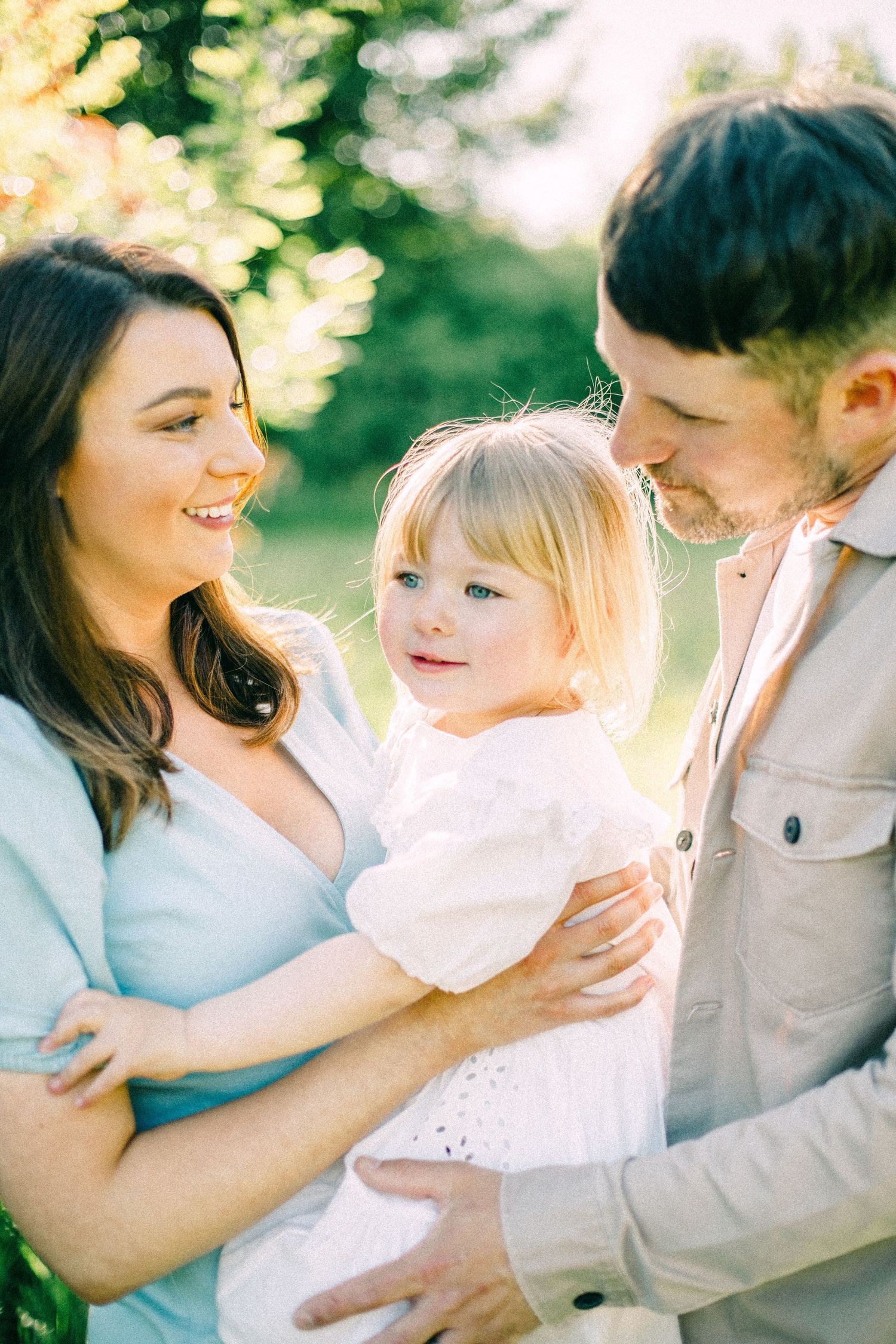 Smiling woman in a light blue top and bearded man in a tan shirt hold a blonde toddler in white outdoors in warm golden sunlight.
