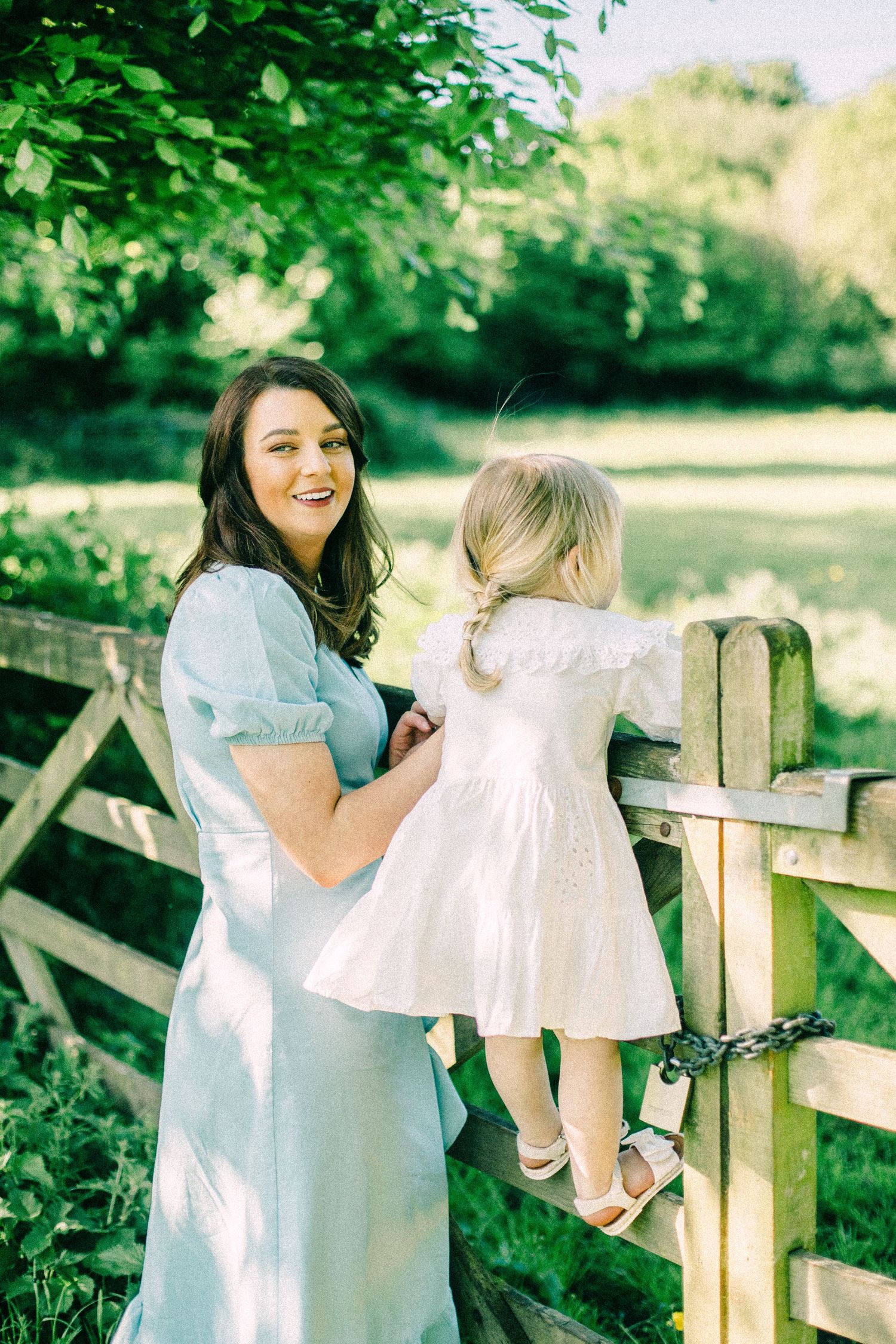 Smiling woman in a light blue dress holds a young girl in a white dress standing on a wooden gate in a green countryside setting.