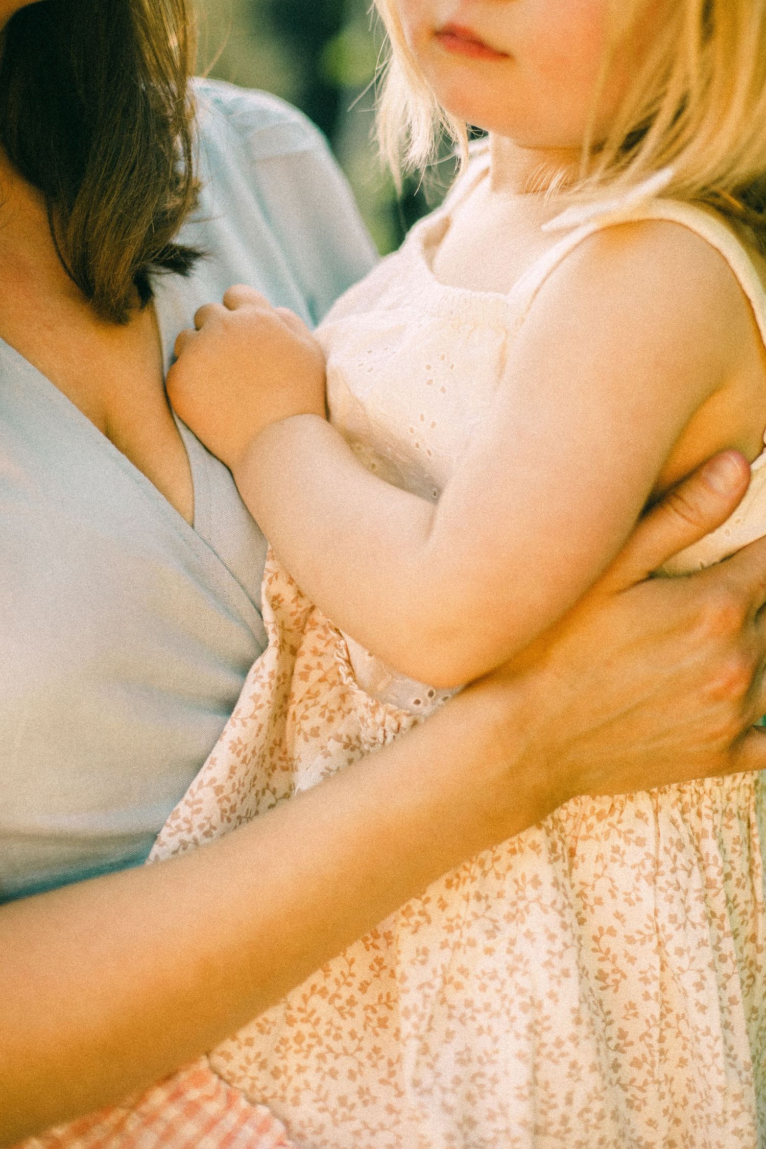 Young blonde girl in a floral sundress being held in the arms of her mother in a soft blue top, bathed in warm natural light.