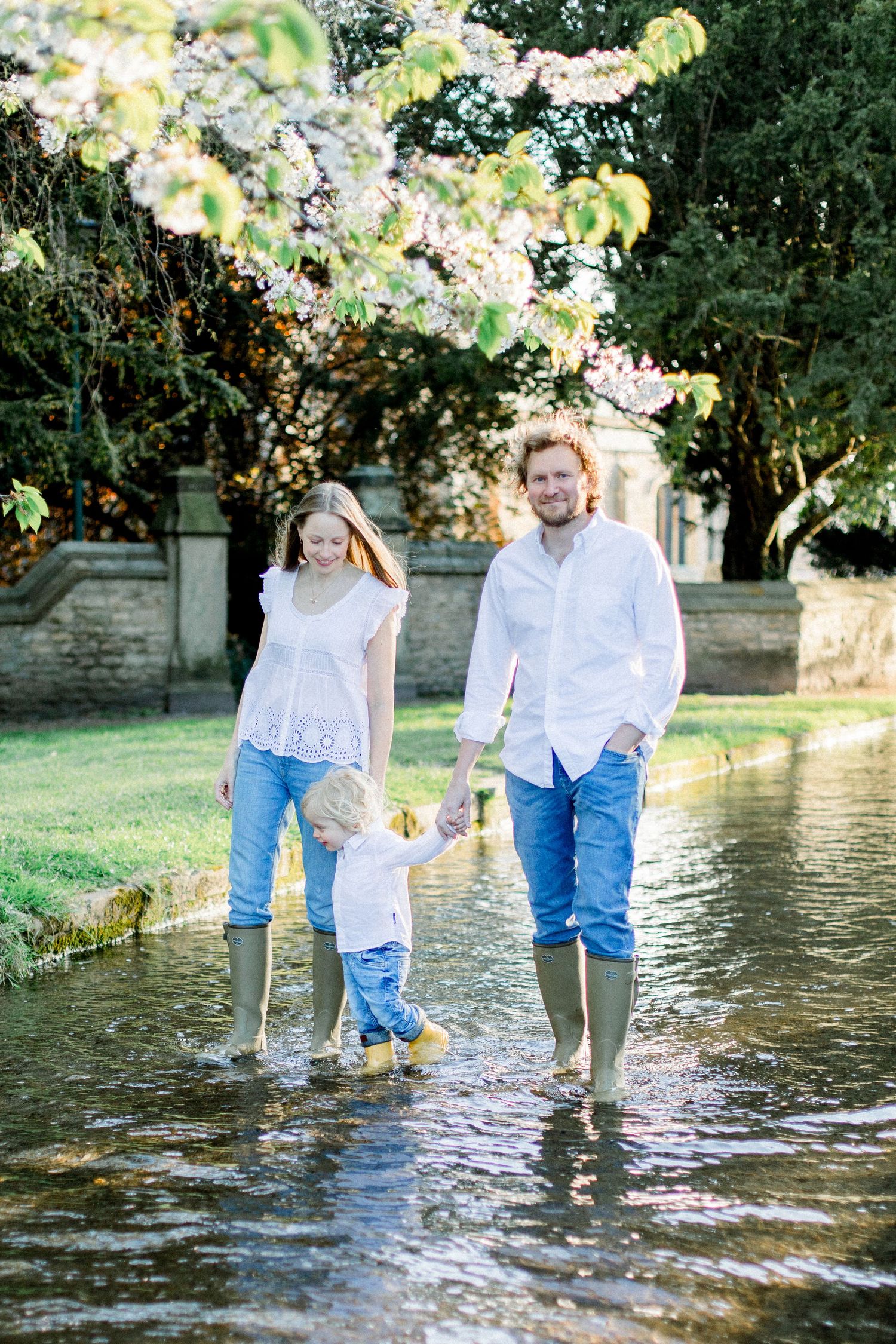A family wearing white and denim stands together in shallow stream water wearing rain boots.