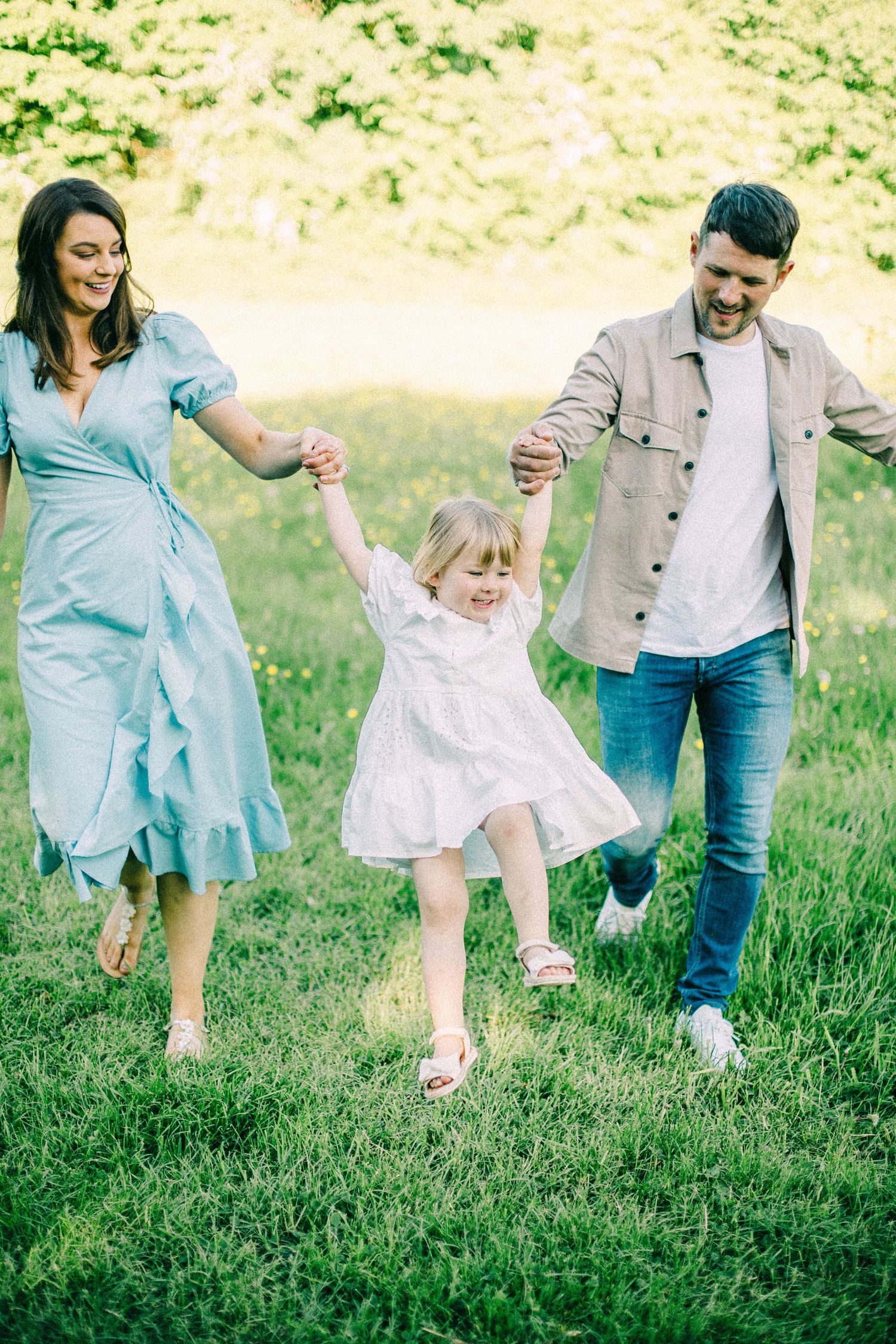 Smiling parents hold hands with a laughing toddler in a white dress as the family walks through a sunny green meadow.