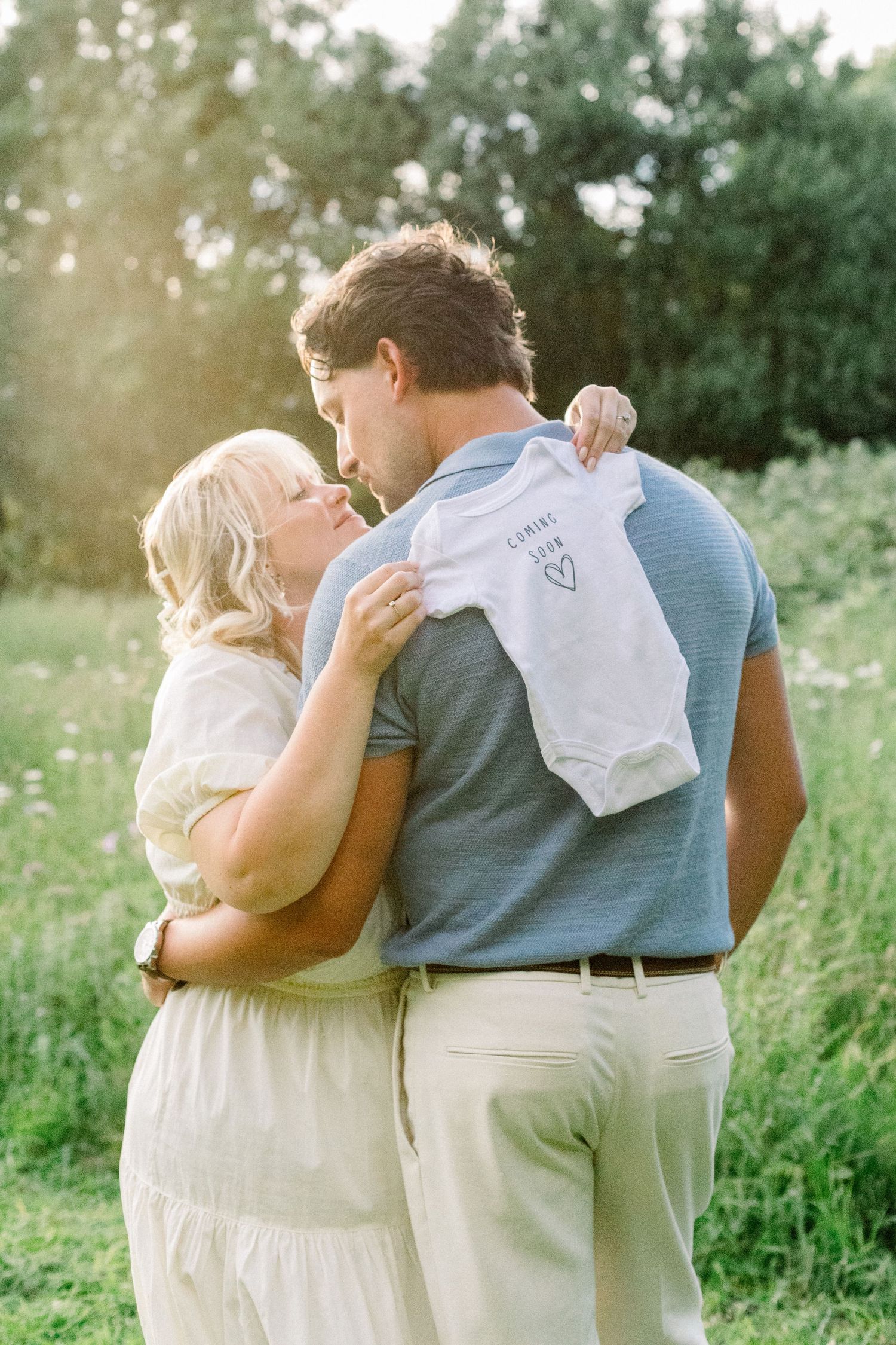 Expecting couple embracing in a sunlit grassy field while holding a white baby onesie with a heart, a sweet pregnancy announcement.