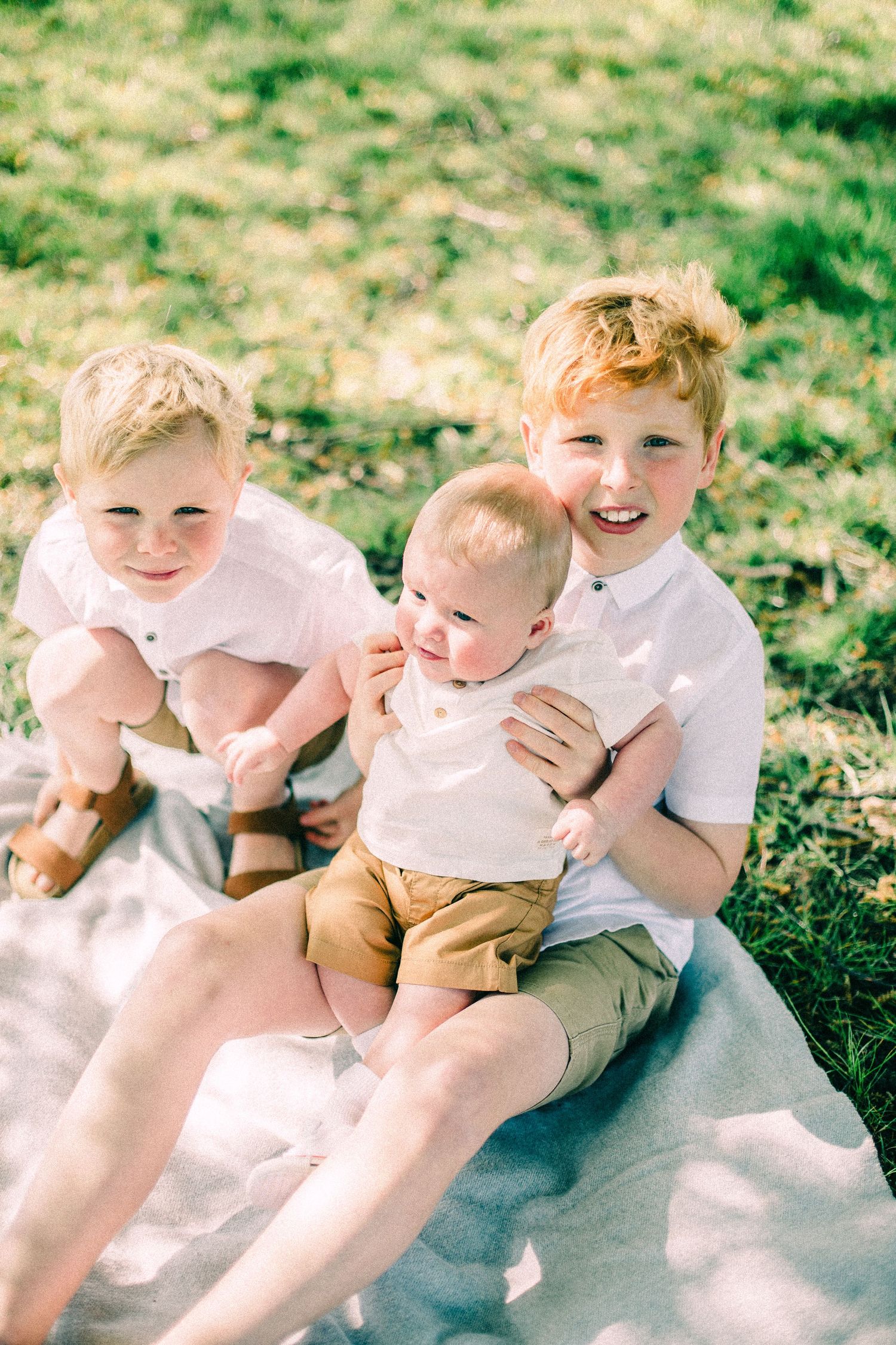 Three smiling young boys in white shirts and tan shorts sit together on a blanket in a sunny grassy park.