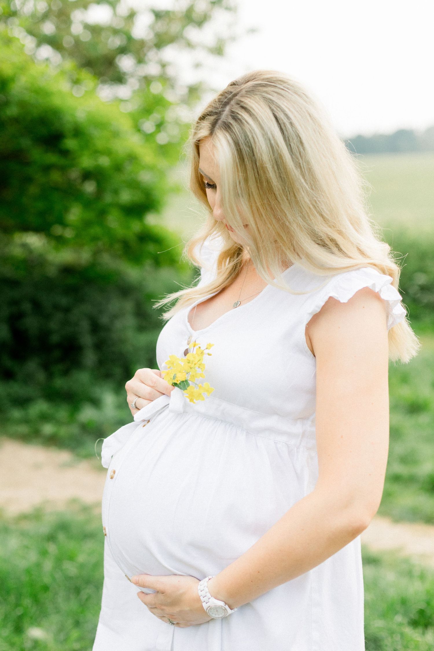 Pregnant woman with long blonde hair in a white dress holds small yellow flowers against her baby bump in a sunny green field.