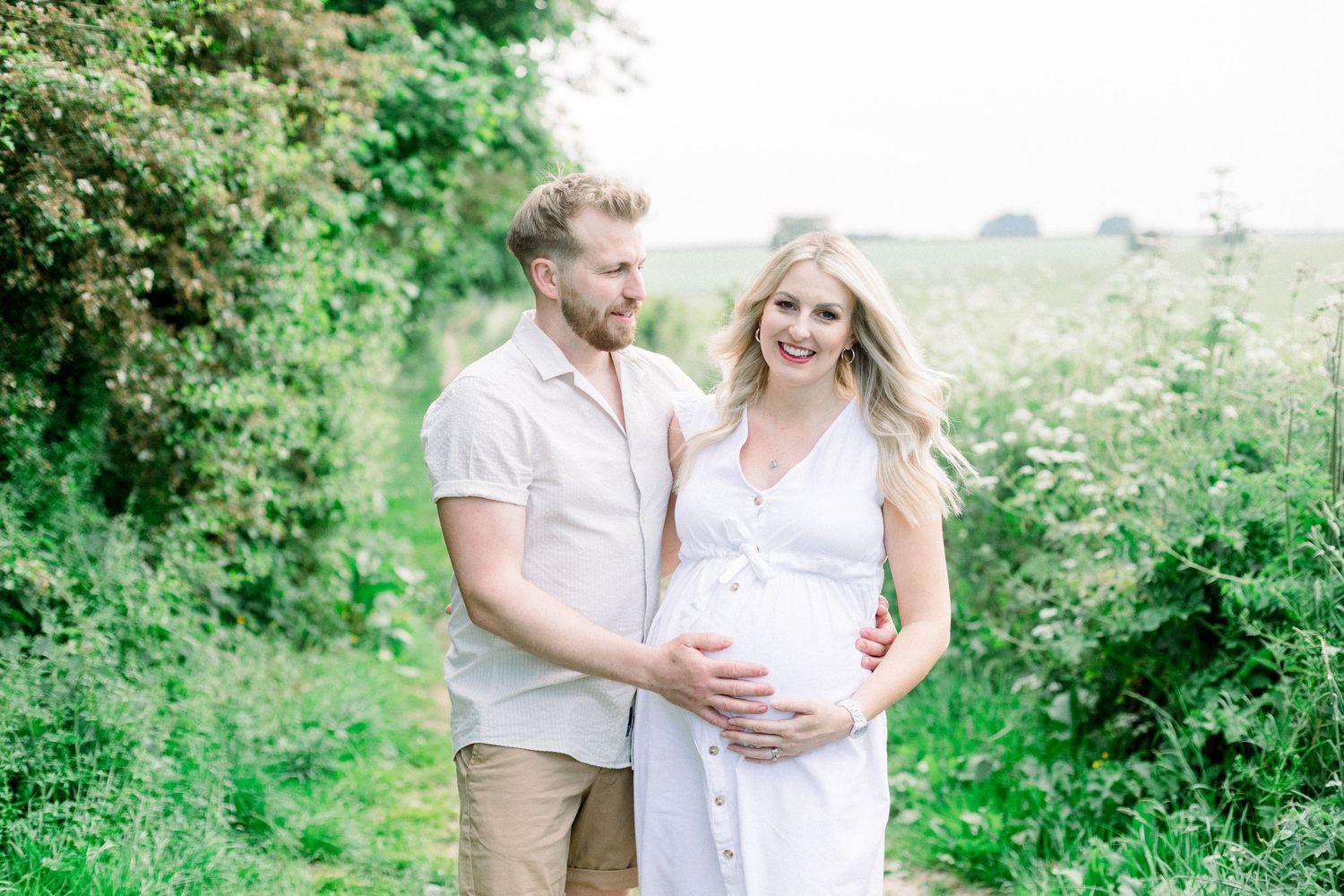 Couple posing outdoors on a garden path, woman in white dress cradling baby bump, man in white shirt embracing her.