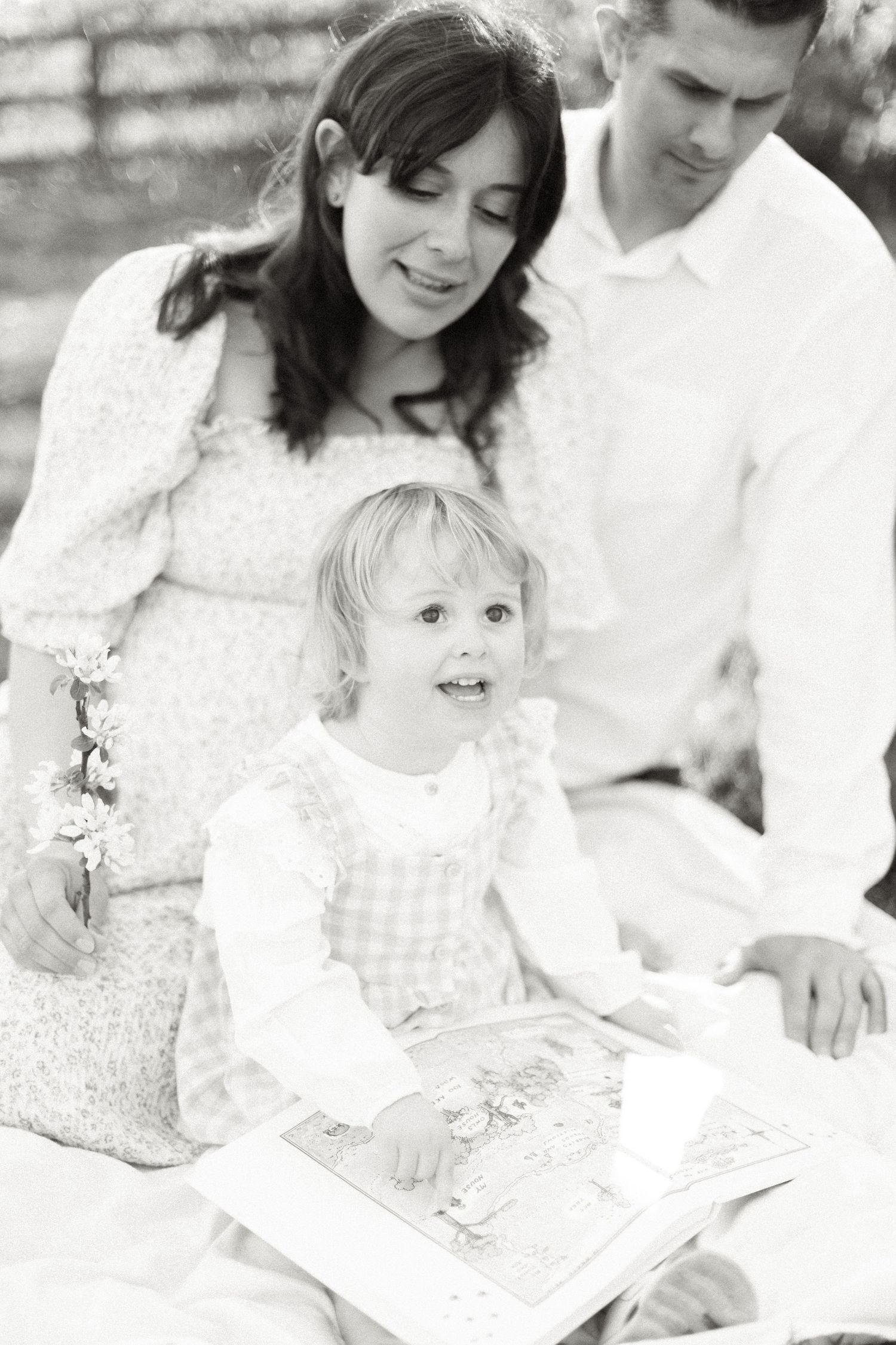 Black and white photo of a pregnant woman and man reading a picture book with their young blonde toddler outdoors.