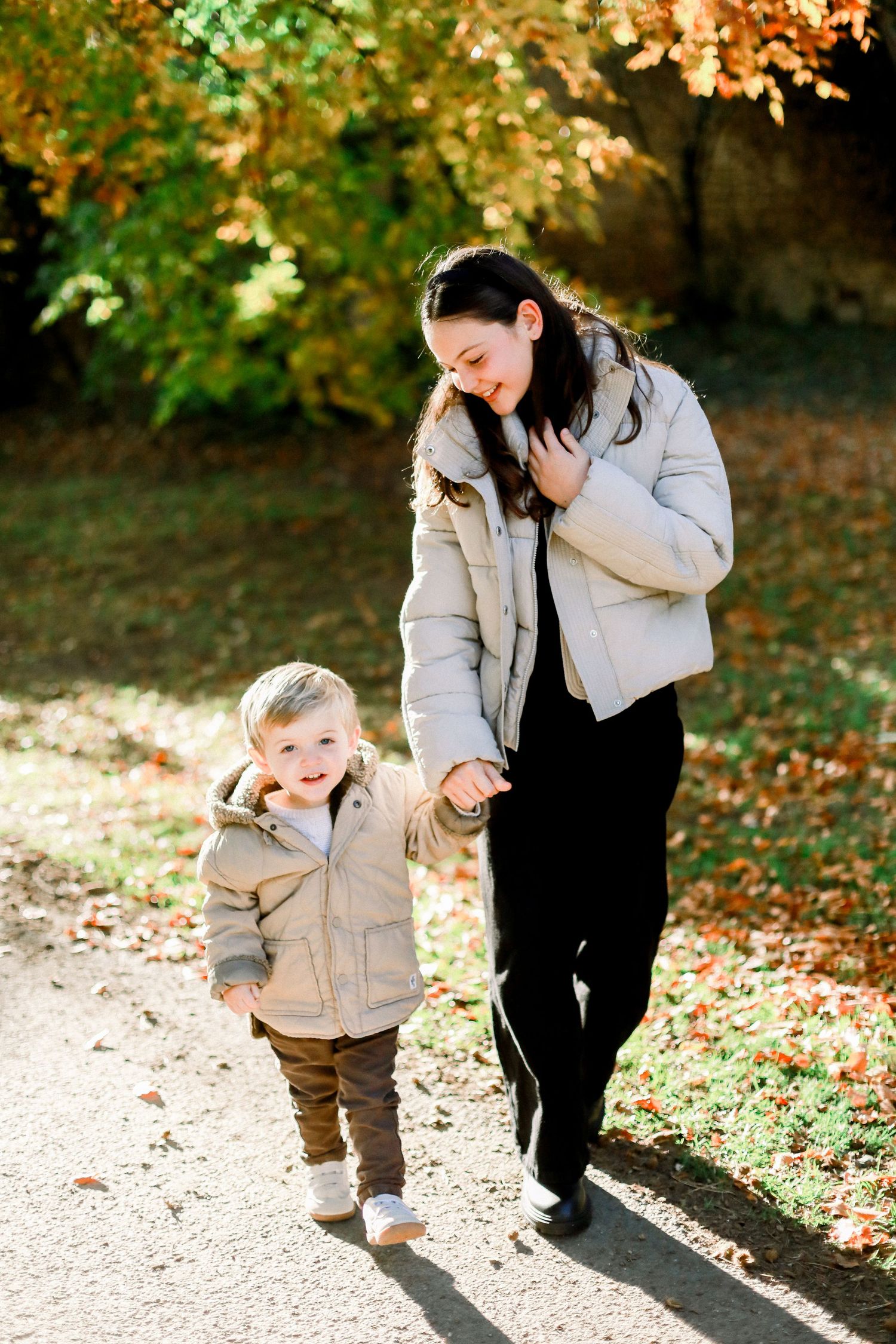 Brother and sister walking hand in hand on autumn park path, both wearing beige jackets with colorful fall foliage.