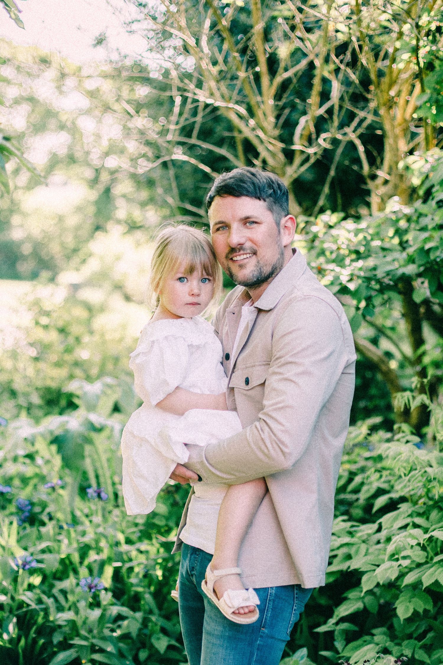 Smiling bearded man in a beige jacket holding a little girl in a white dress amid lush green garden foliage and purple flowers.
