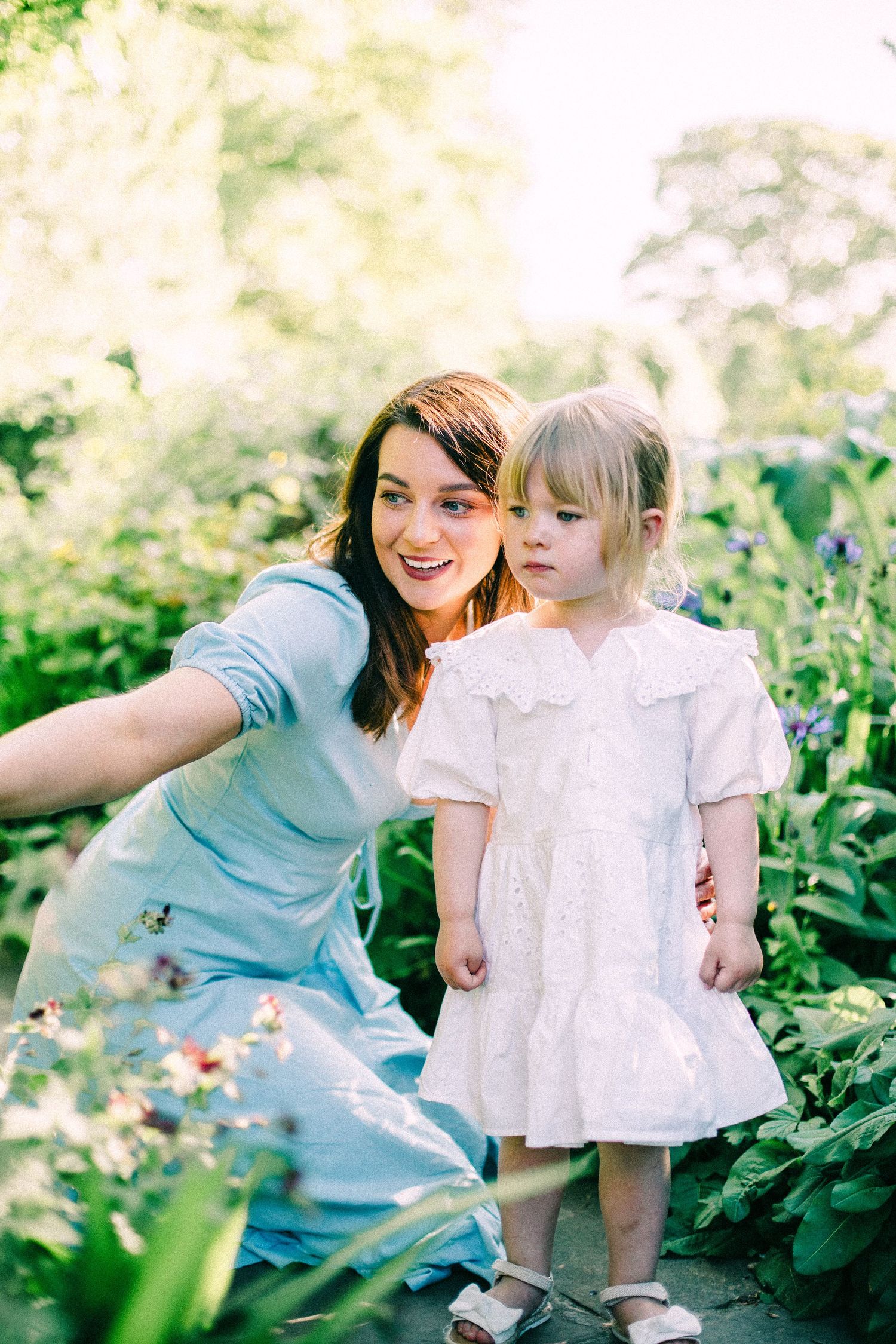 Smiling woman in a blue dress crouches beside a toddler in a white lace dress among lush green garden plants and flowers.