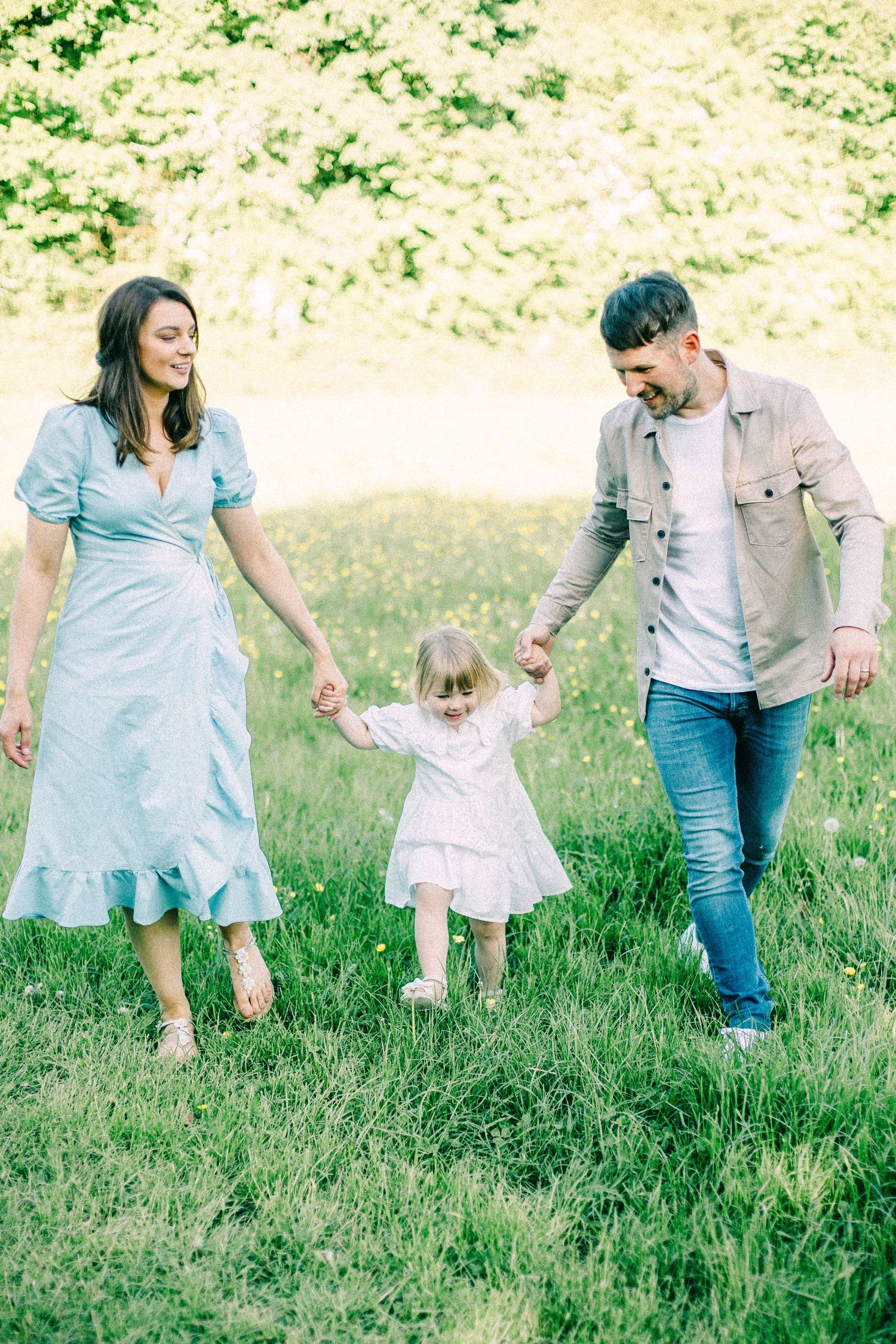 A smiling couple holds hands with their toddler daughter as they walk together through a sunny green meadow.