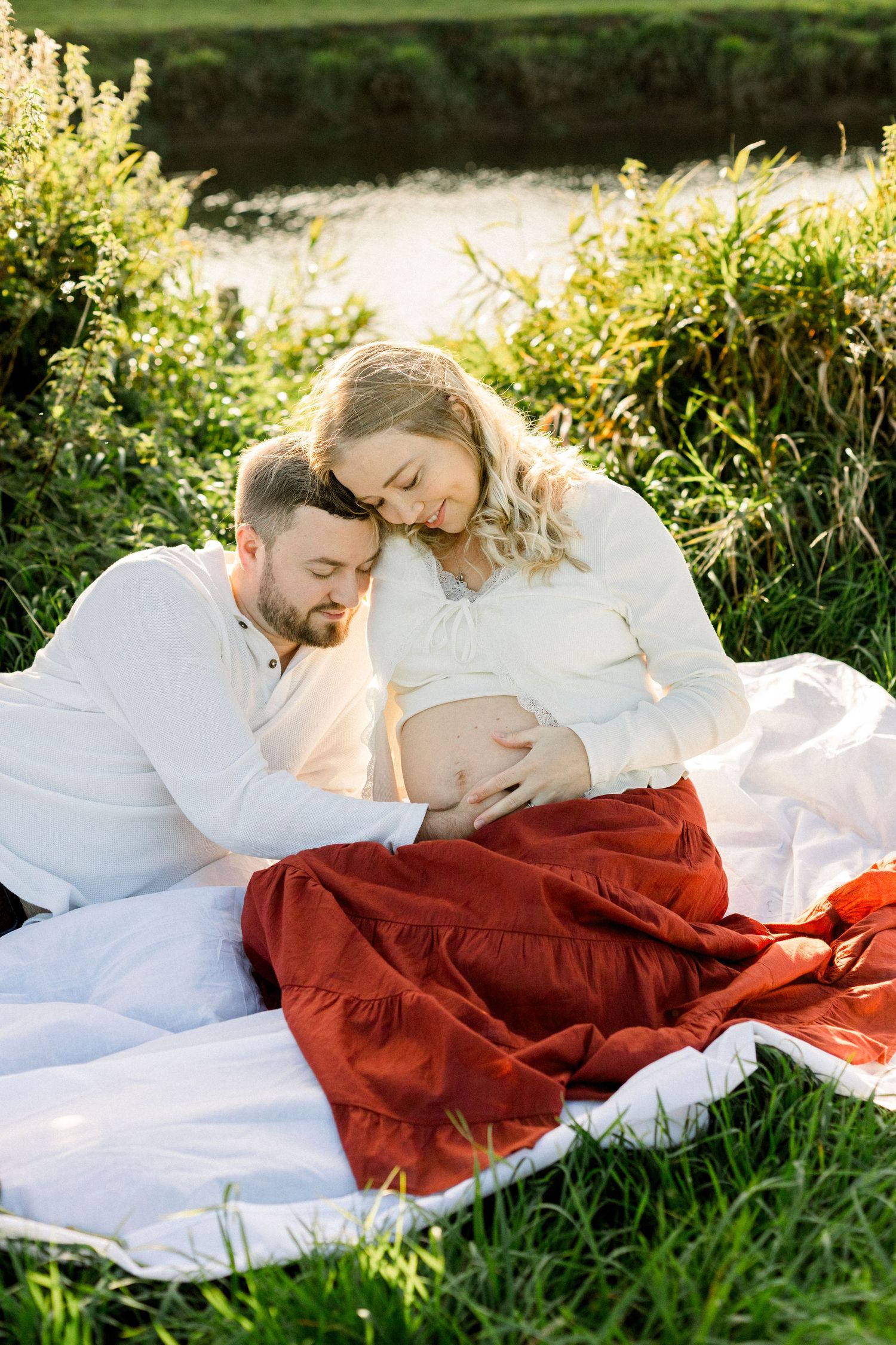 Expectant couple relaxing on a white blanket by a sunlit river, the man resting his head on the pregnant woman's belly in a rust skirt.