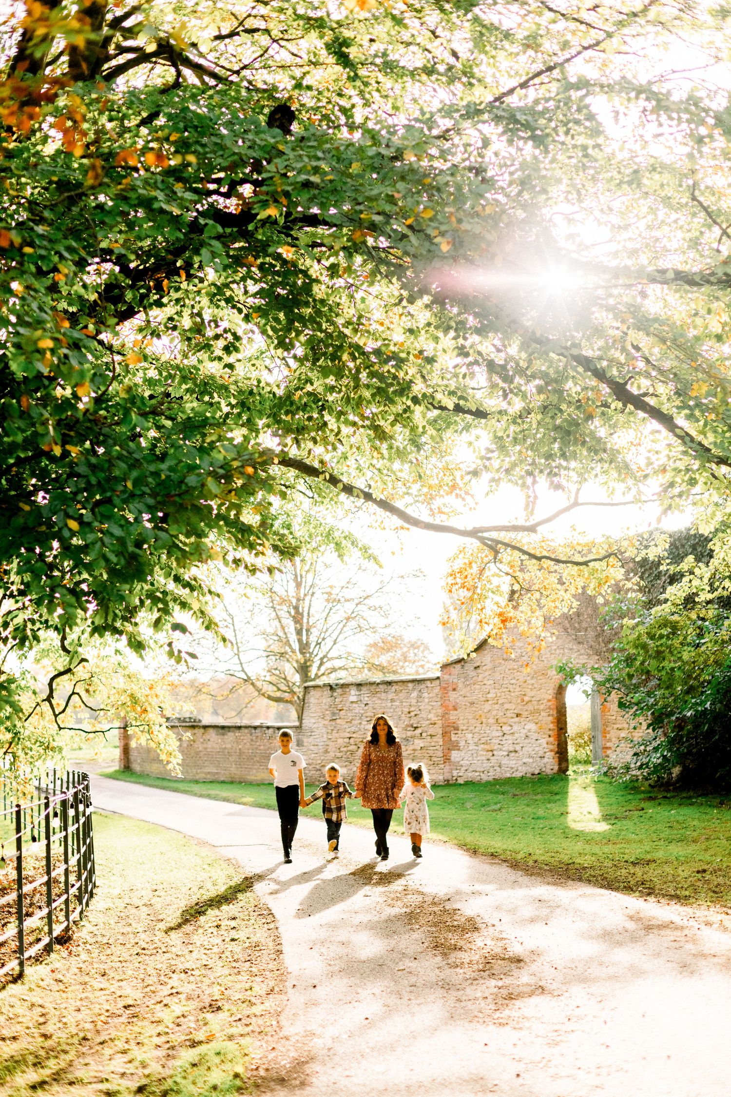 People walking along a misty autumn path with stone walls and golden leaves beneath sunlit trees.