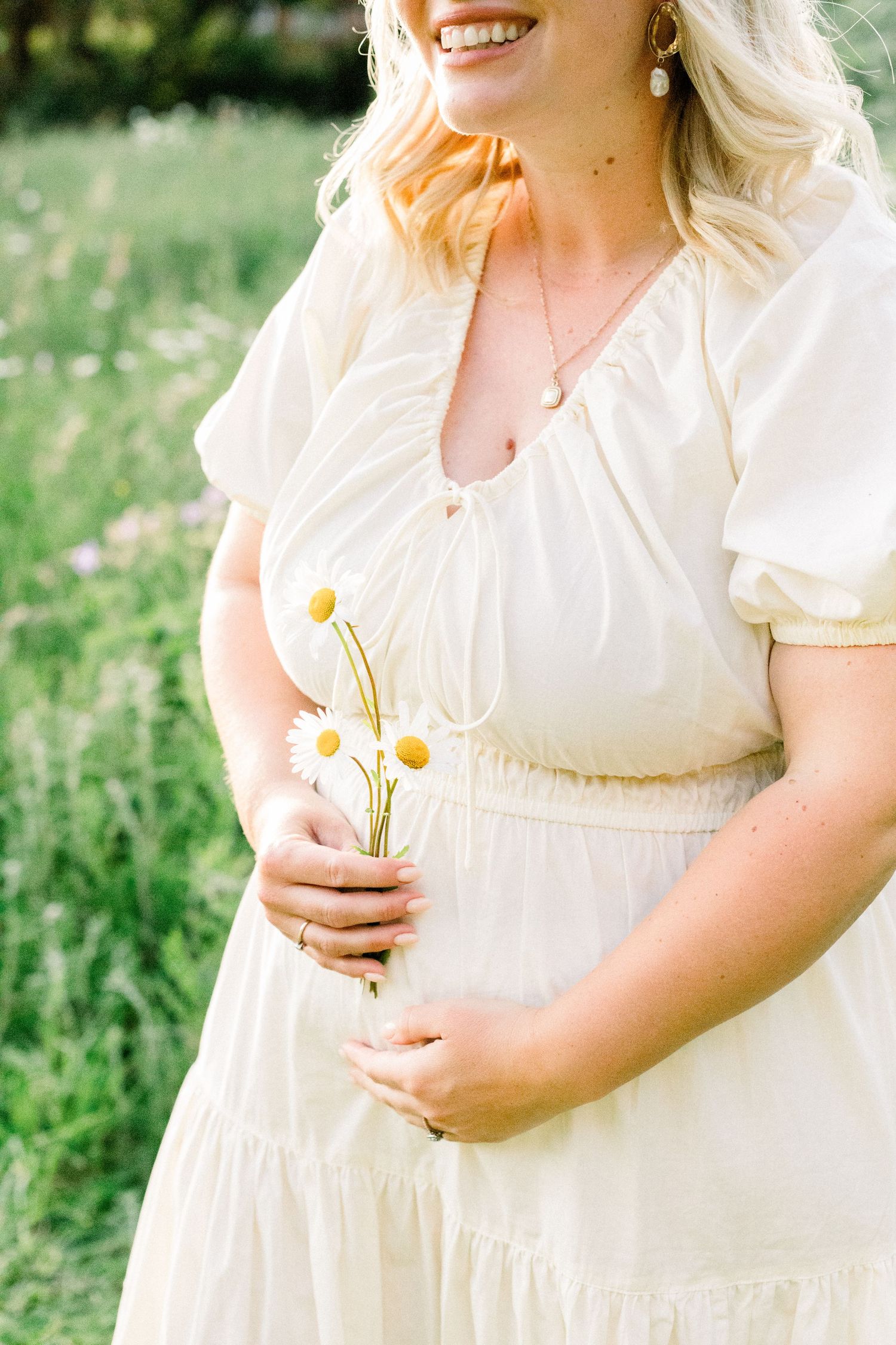 Smiling blonde woman in cream dress holding white daisies outdoors in a green field.