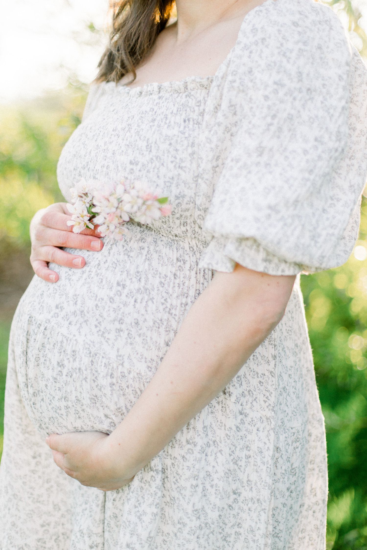 Pregnant woman in floral dress cradling baby bump while holding pink spring blossoms outdoors in soft sunlight.