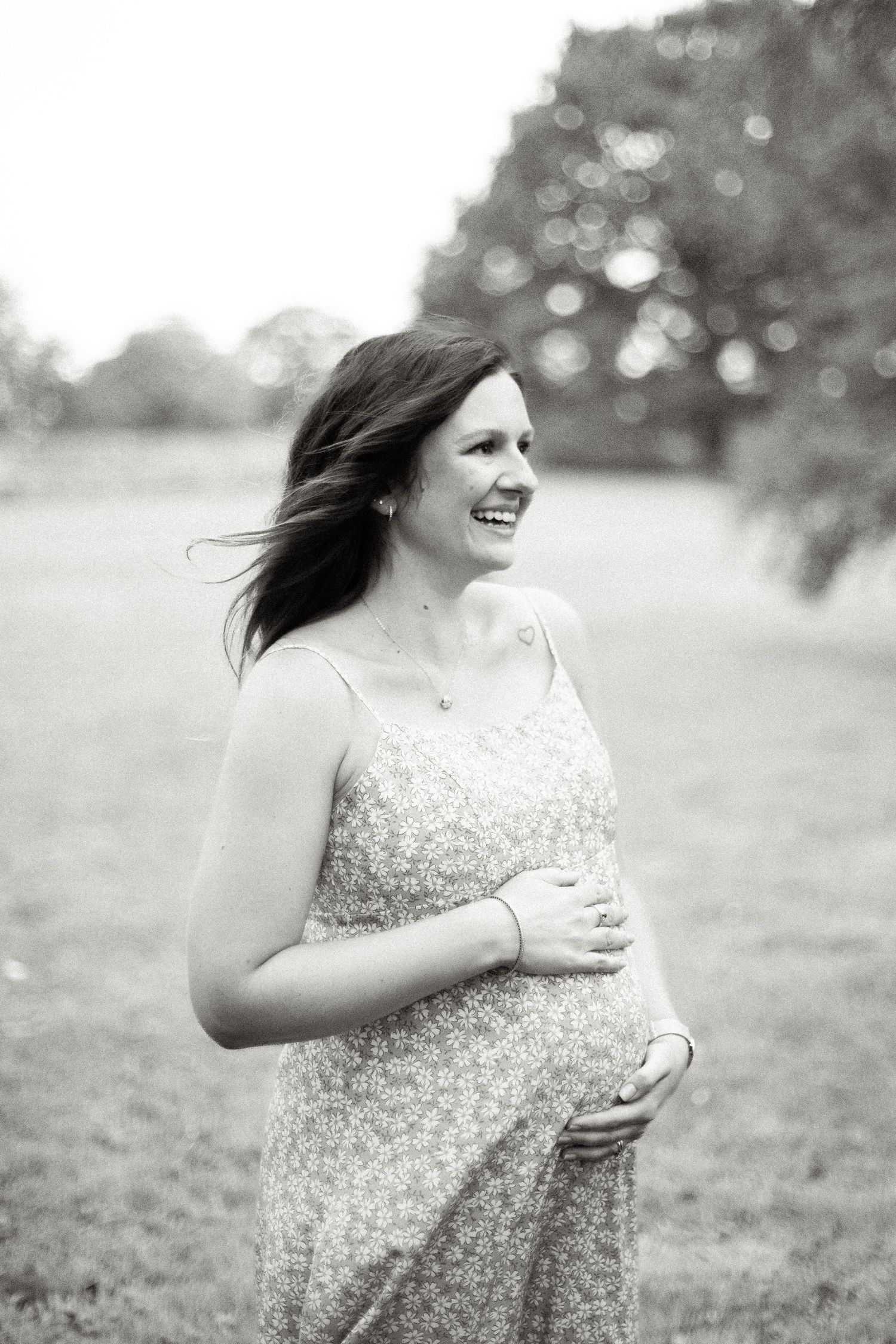 Black and white maternity photo of smiling pregnant woman in floral dress holding belly outdoors in park.