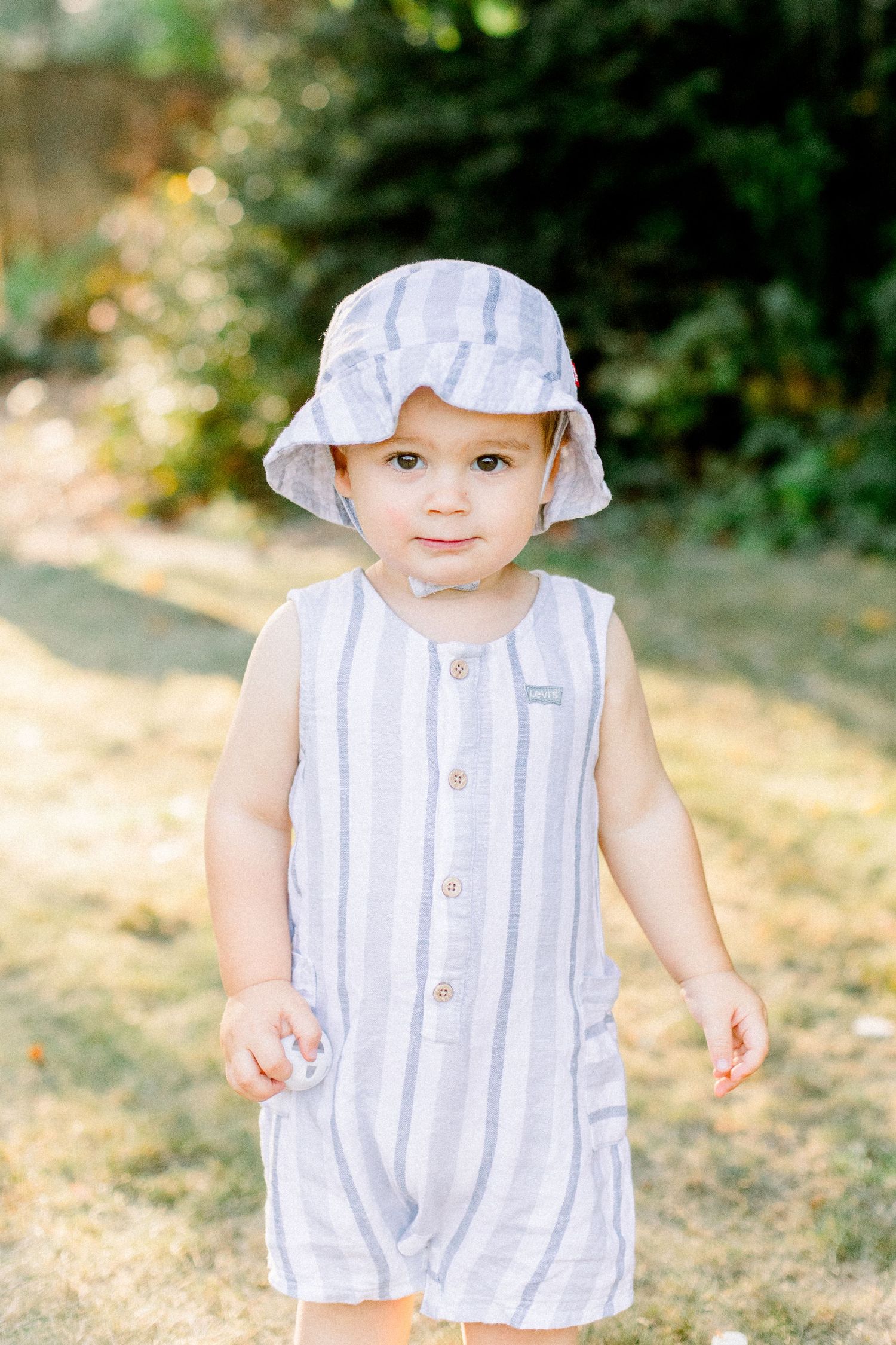 A young child wearing a striped summer romper and sun hat plays in a sunny garden setting.
