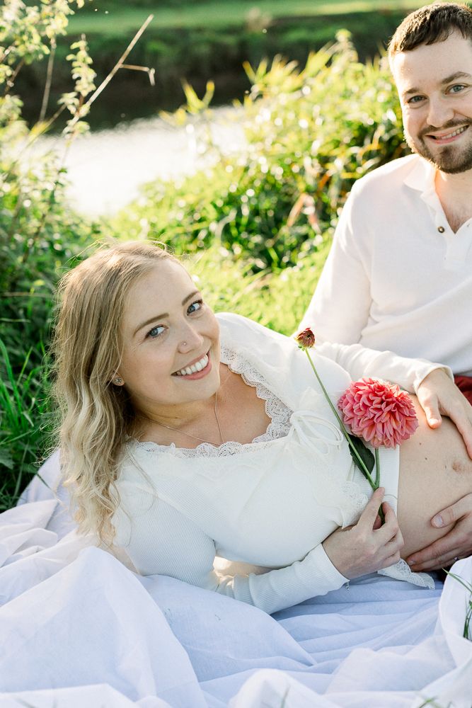 Smiling pregnant woman in white dress holding pink flower with partner outdoors by greenery.