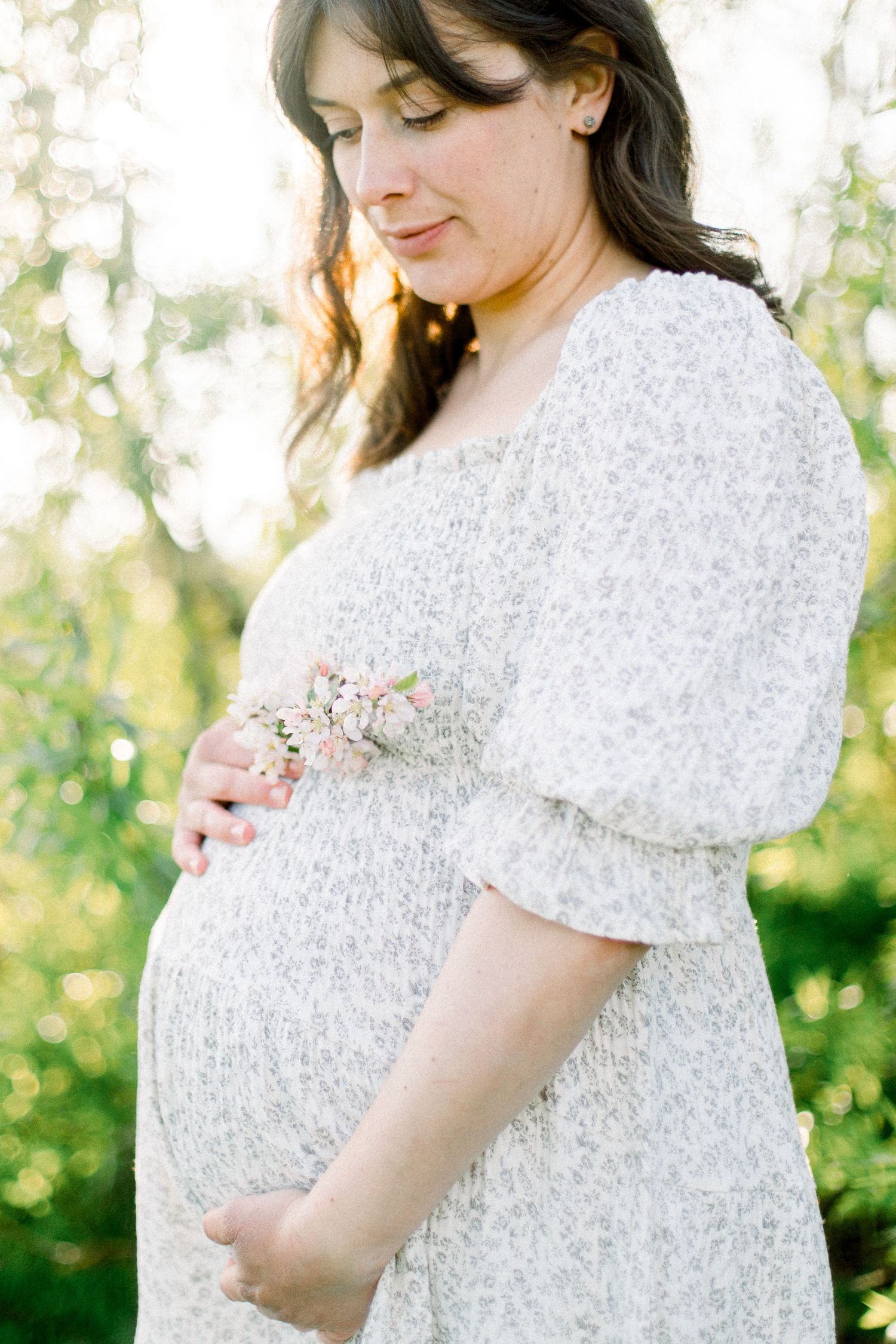 Pregnant woman in white floral dress holding small pink flowers, cradling baby bump outdoors in soft natural light.