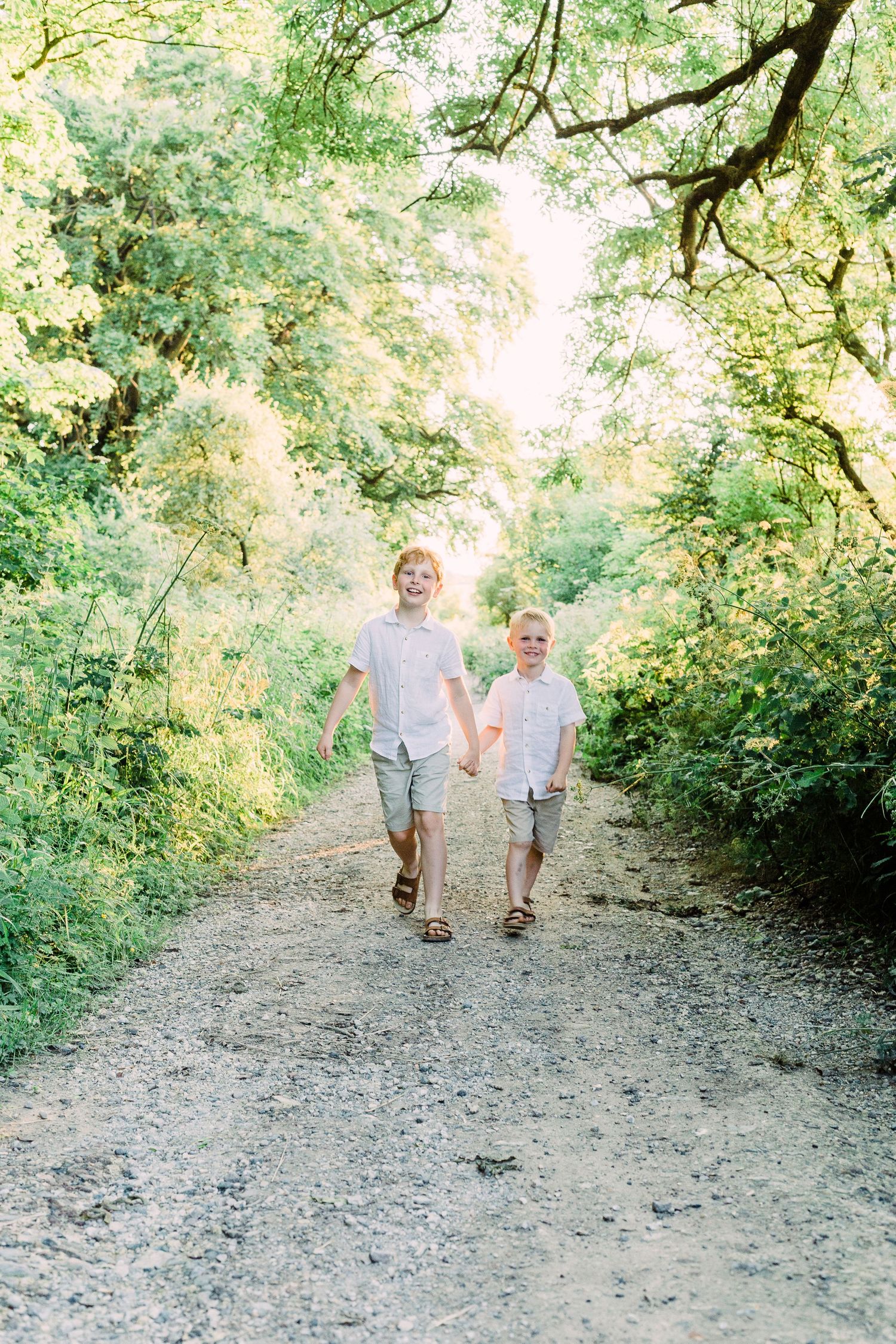 Two smiling boys in matching white shirts and shorts hold hands walking down a sunlit tree-lined dirt path.
