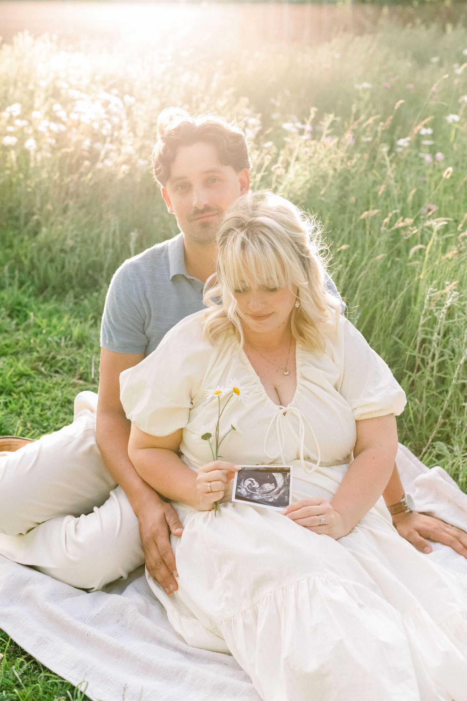 Smiling couple sitting on a white blanket in a sunlit wildflower meadow, holding an ultrasound photo during a maternity photoshoot.
