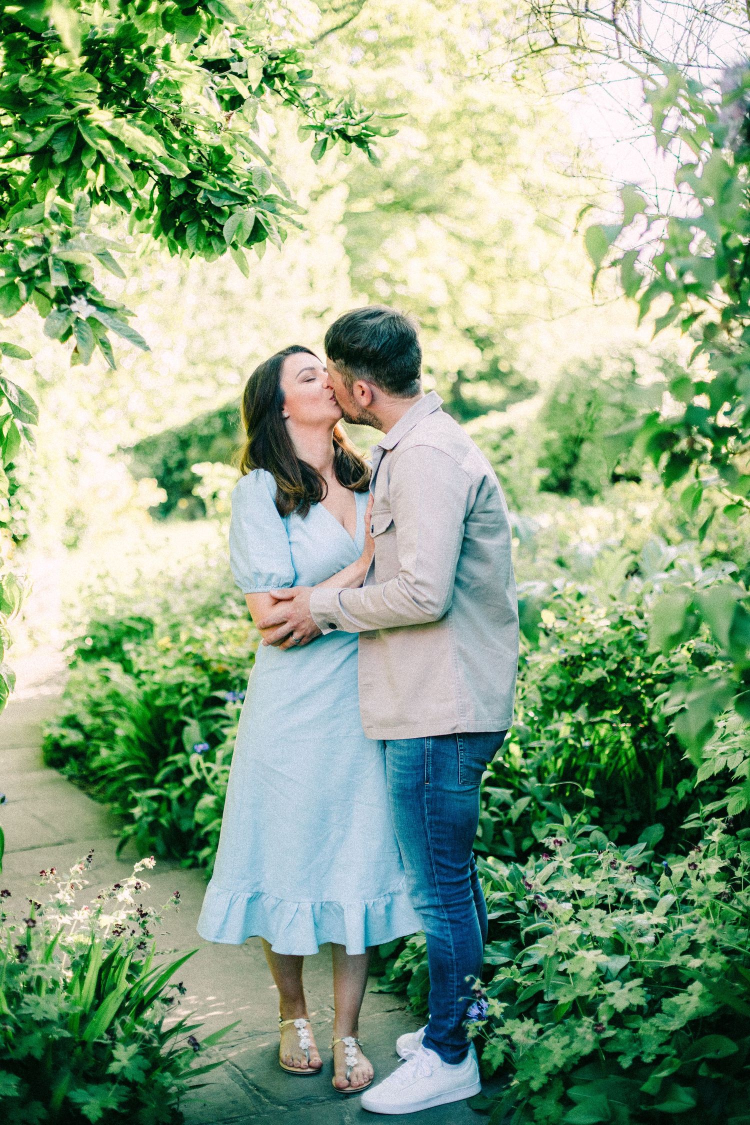 Couple in a light blue dress and beige jacket share a kiss on a stone path surrounded by lush green garden foliage.