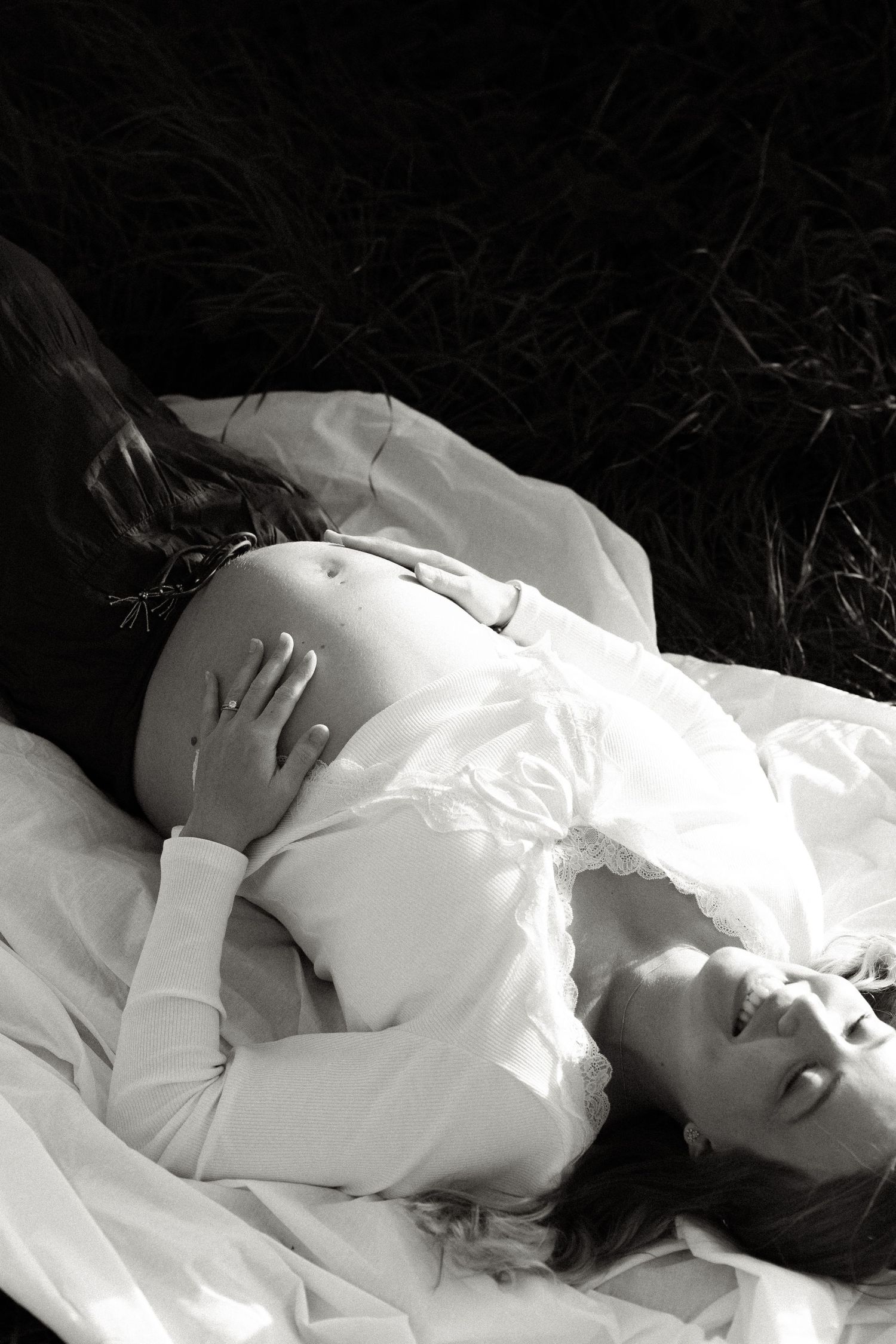 Black and white photo of a smiling pregnant woman lying on white fabric outdoors, gently cradling her bare belly.