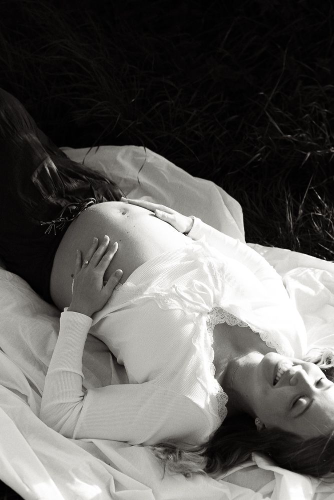 Black and white photo of a pregnant woman lying on white fabric outdoors, smiling peacefully with hands on belly.