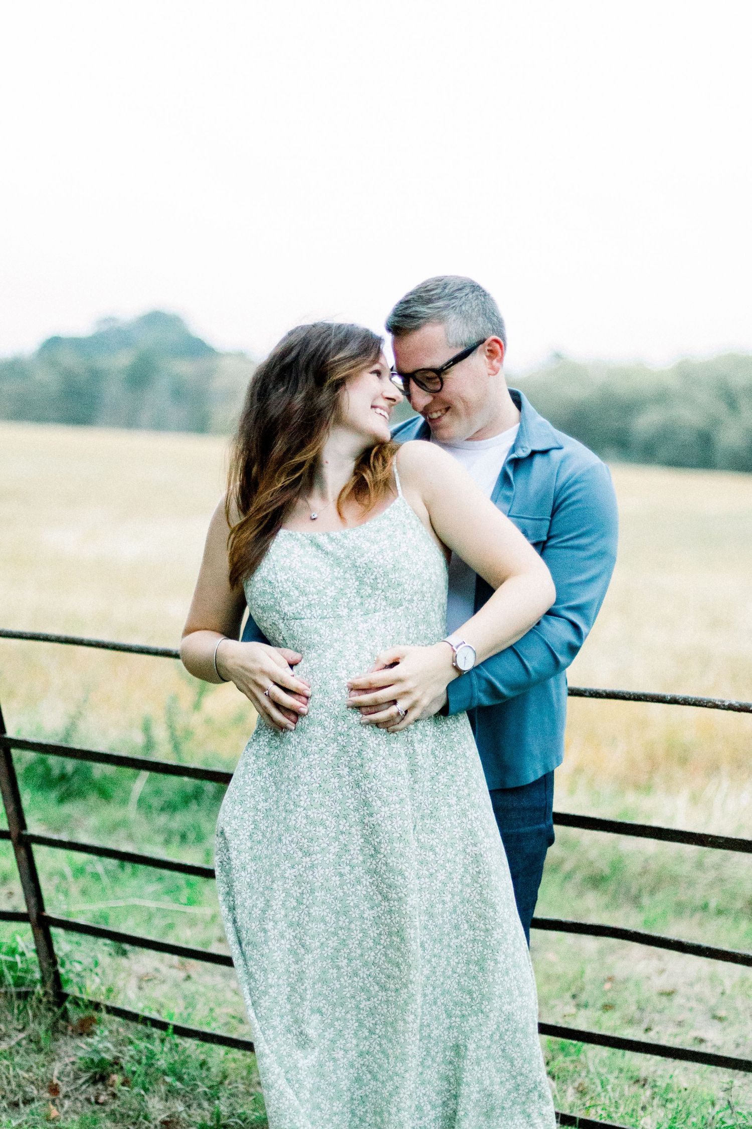 A couple shares a tender moment by a rustic fence in a field, the woman in a floral dress smiling as the man embraces her.
