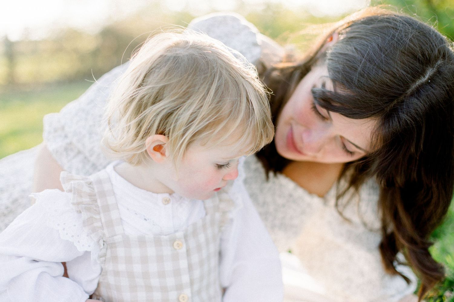 A mother with dark hair tenderly gazes at her blonde toddler in soft natural light outdoors.