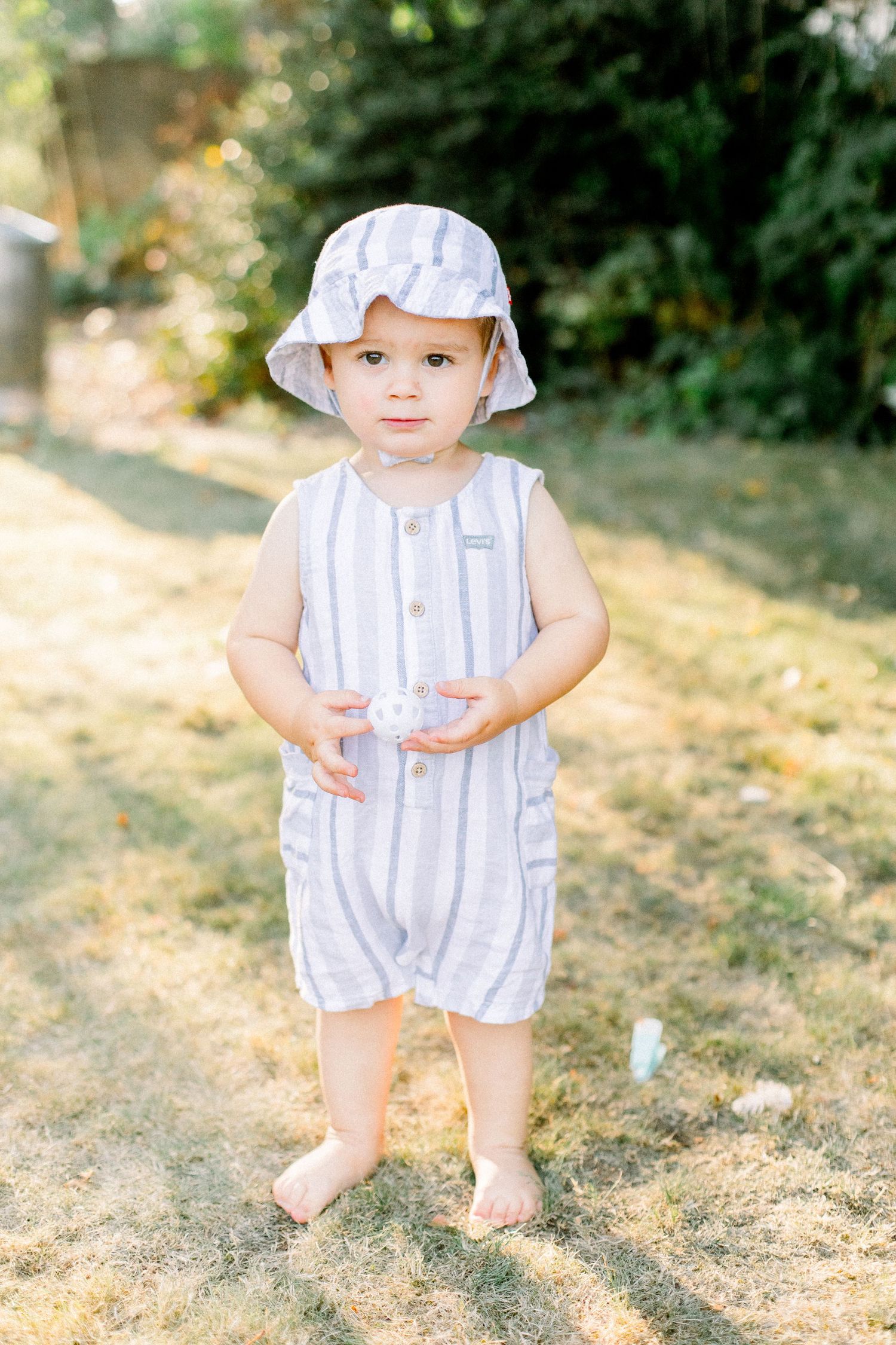 Child in white striped romper and sun hat standing barefoot on dry grass.