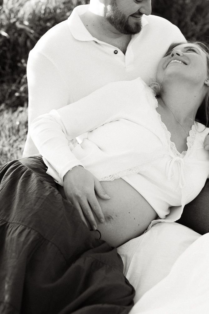 Black and white photo of a couple outdoors, man tenderly holding pregnant womans belly as she leans back smiling.