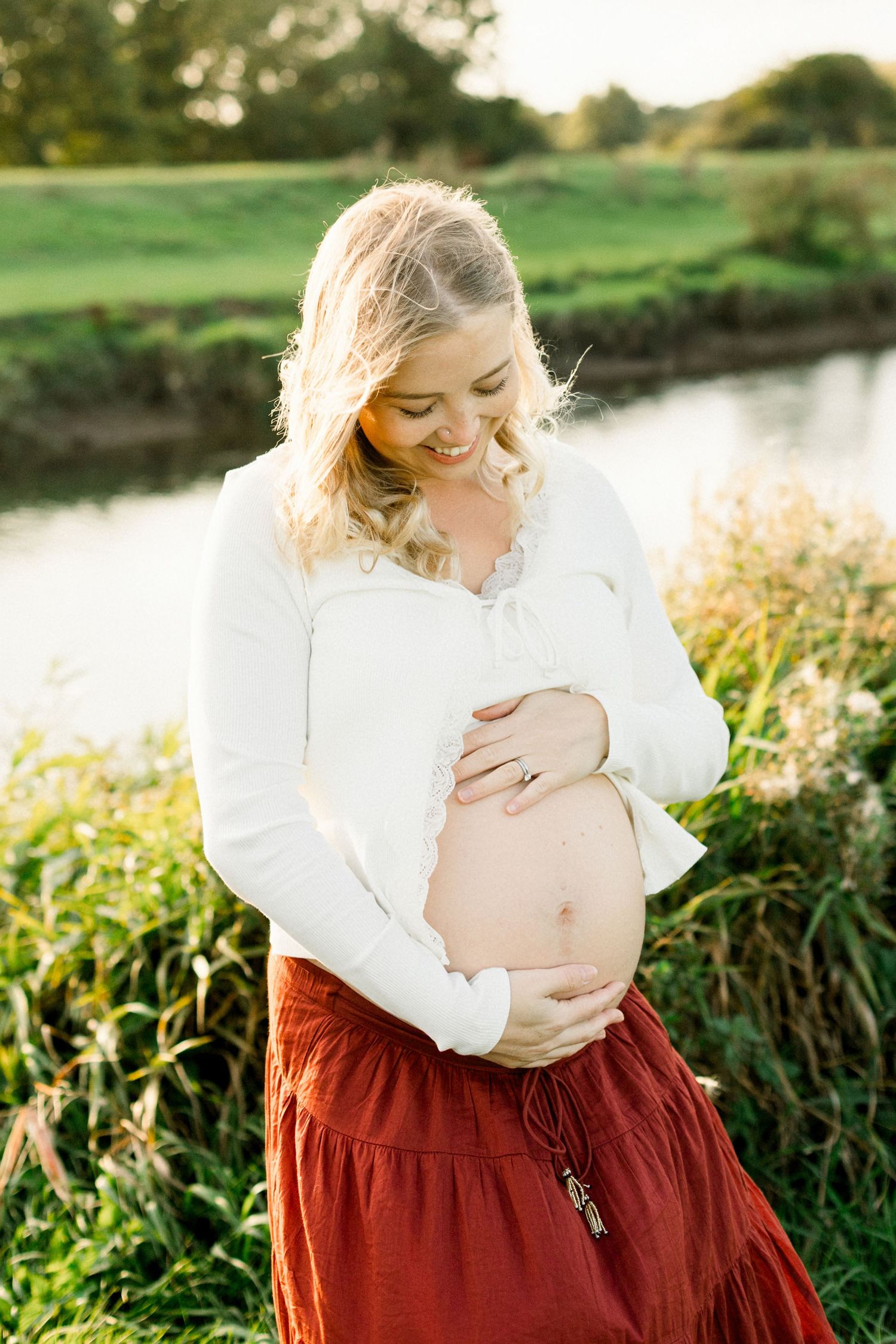 Smiling pregnant woman in white top and red skirt cradling her baby bump outdoors near a serene pond.