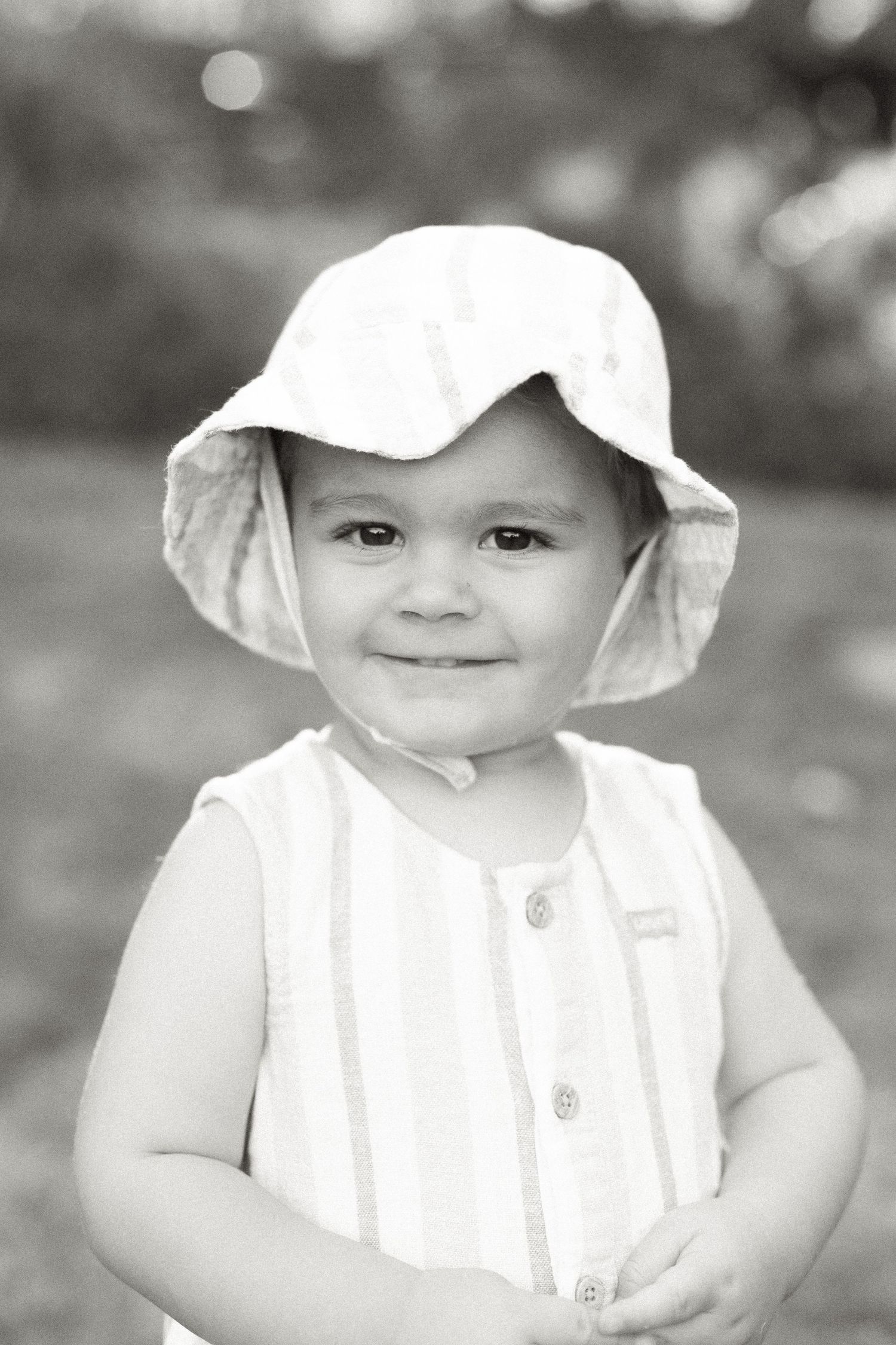 Black and white portrait of a child in a white sun hat and button-up romper.