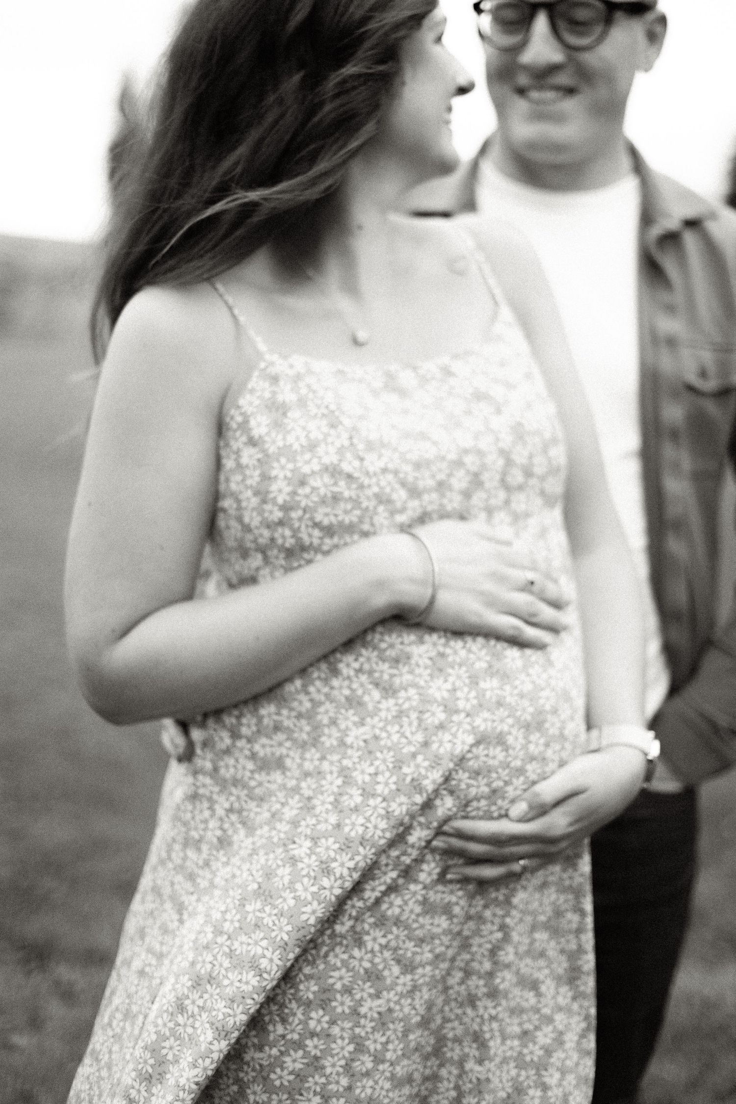 Black and white photo of a pregnant woman in floral dress cradling her baby bump with her partner outdoors.