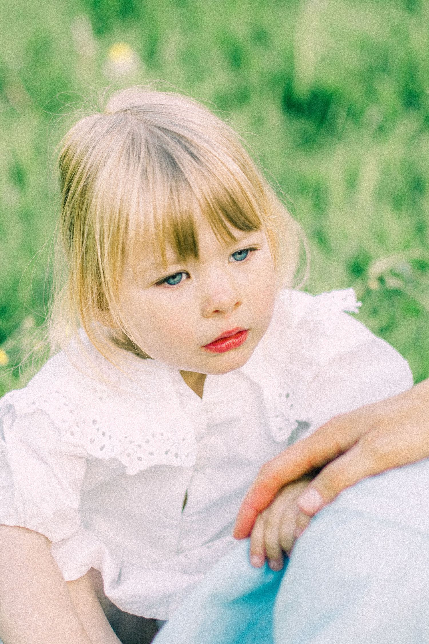 Young blonde girl with bangs and blue eyes in a white lace blouse sits on a green grass meadow as an adult hand rests on her.