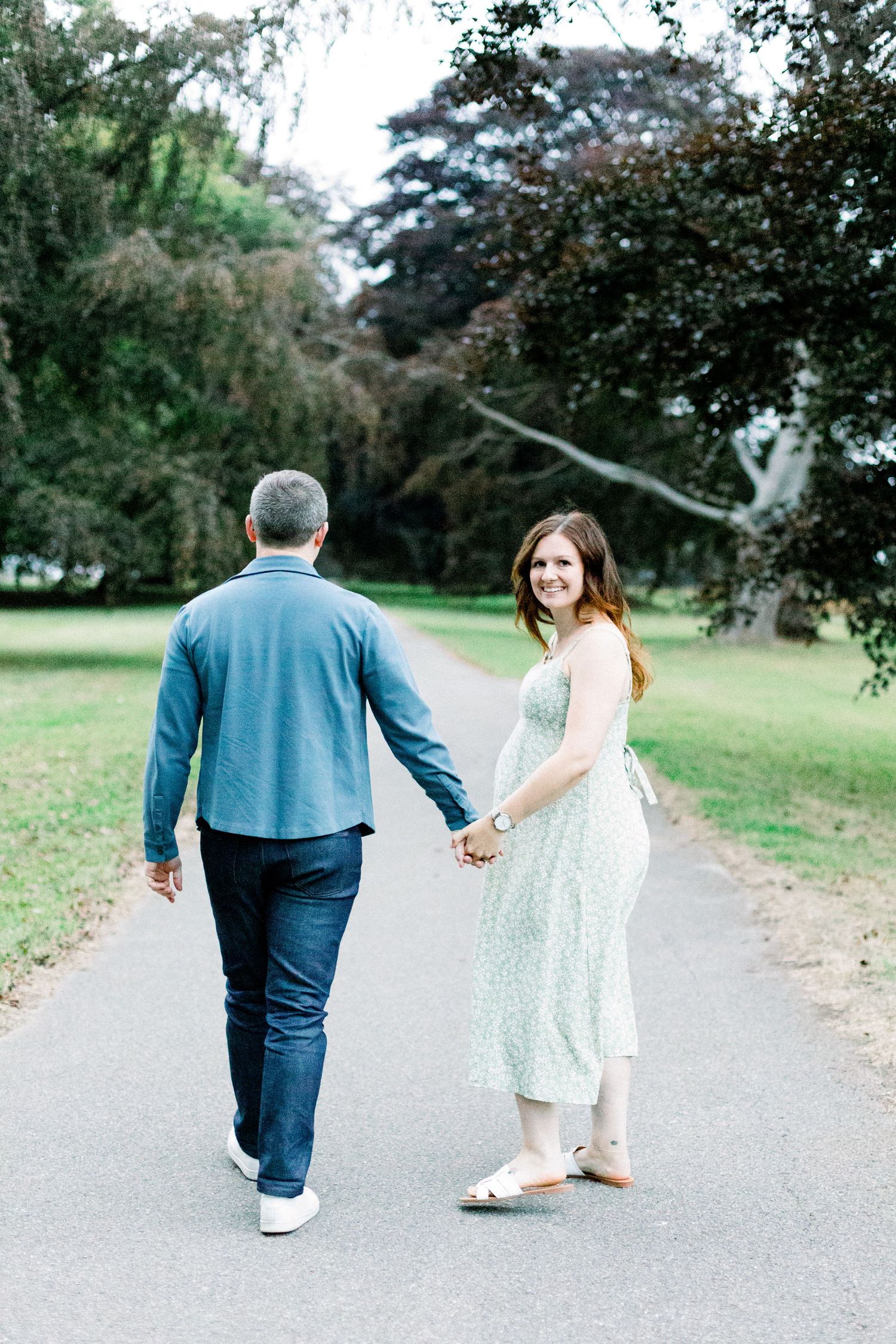 Couple holding hands walking down a park path, woman in floral dress smiling back at camera.