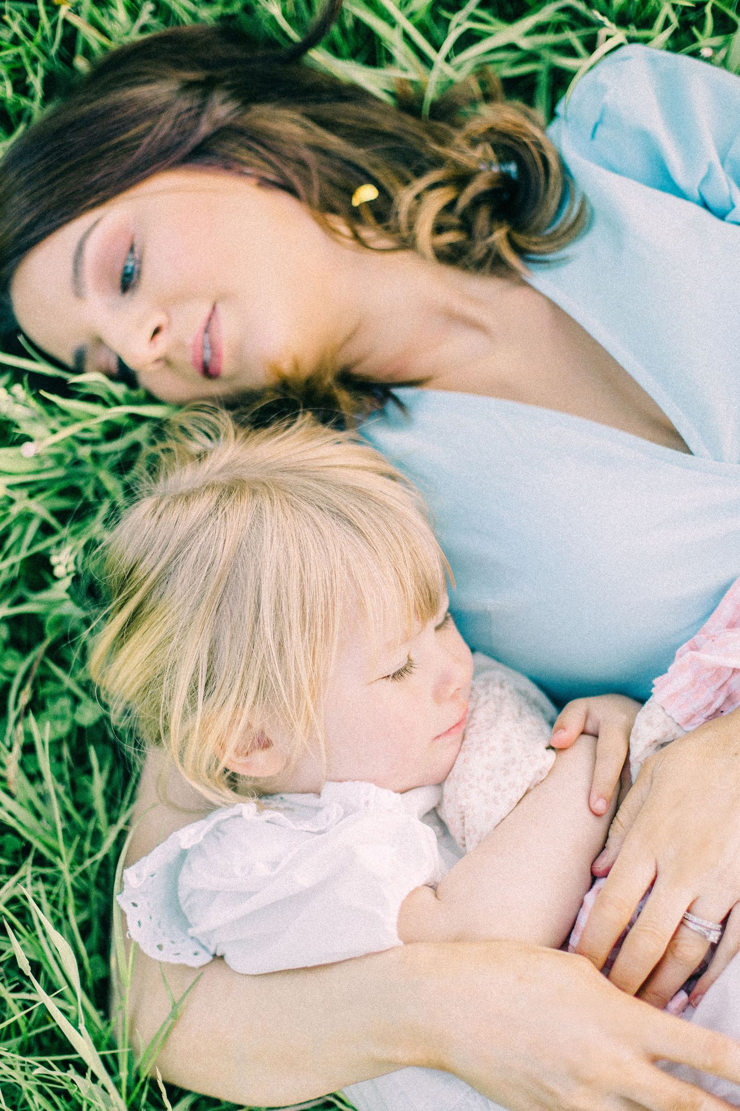 Mother in a light blue top lies in tall green grass cuddling her sleeping blonde toddler in a white lace outfit.