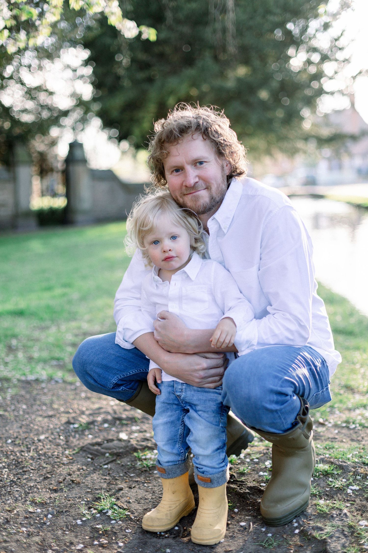 A curly-haired man crouching outdoors hugging a blonde toddler in white shirts, jeans, and yellow boots in a green park.