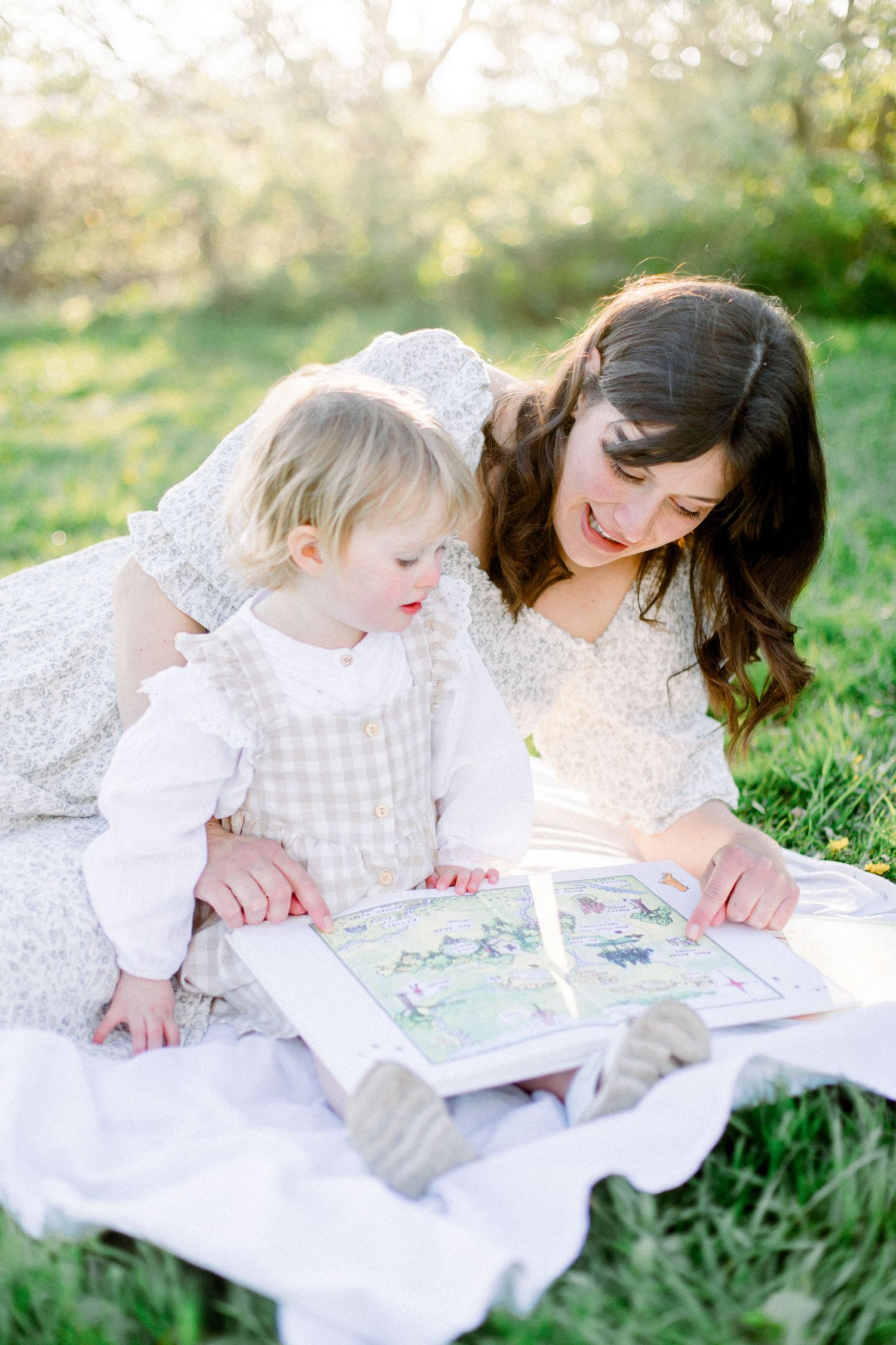 A smiling mother in a floral dress points to pages in a picture book while sitting with her toddler outdoors.