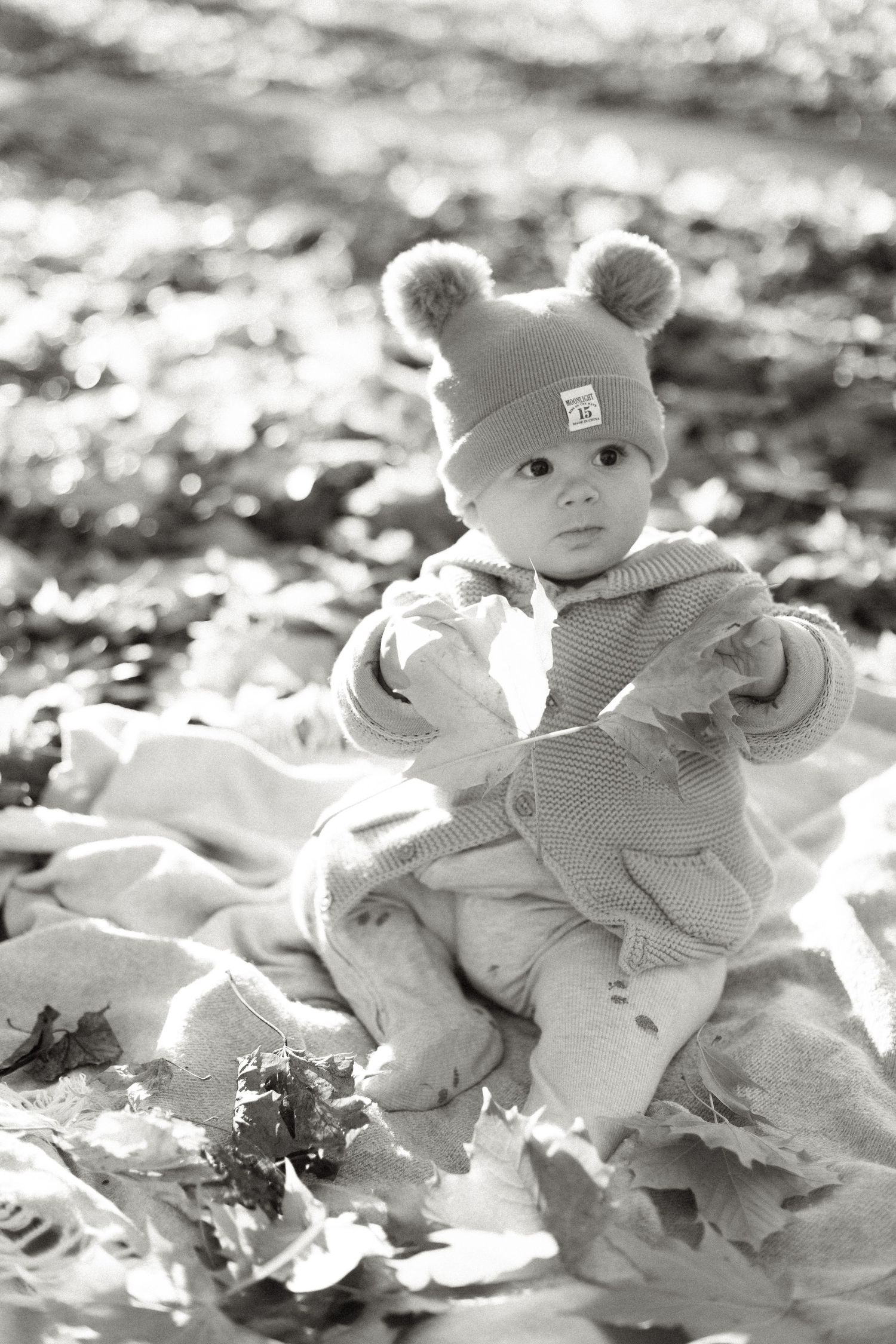 Black and white autumn photo of a baby wearing a bear hat and sweater among fallen leaves.